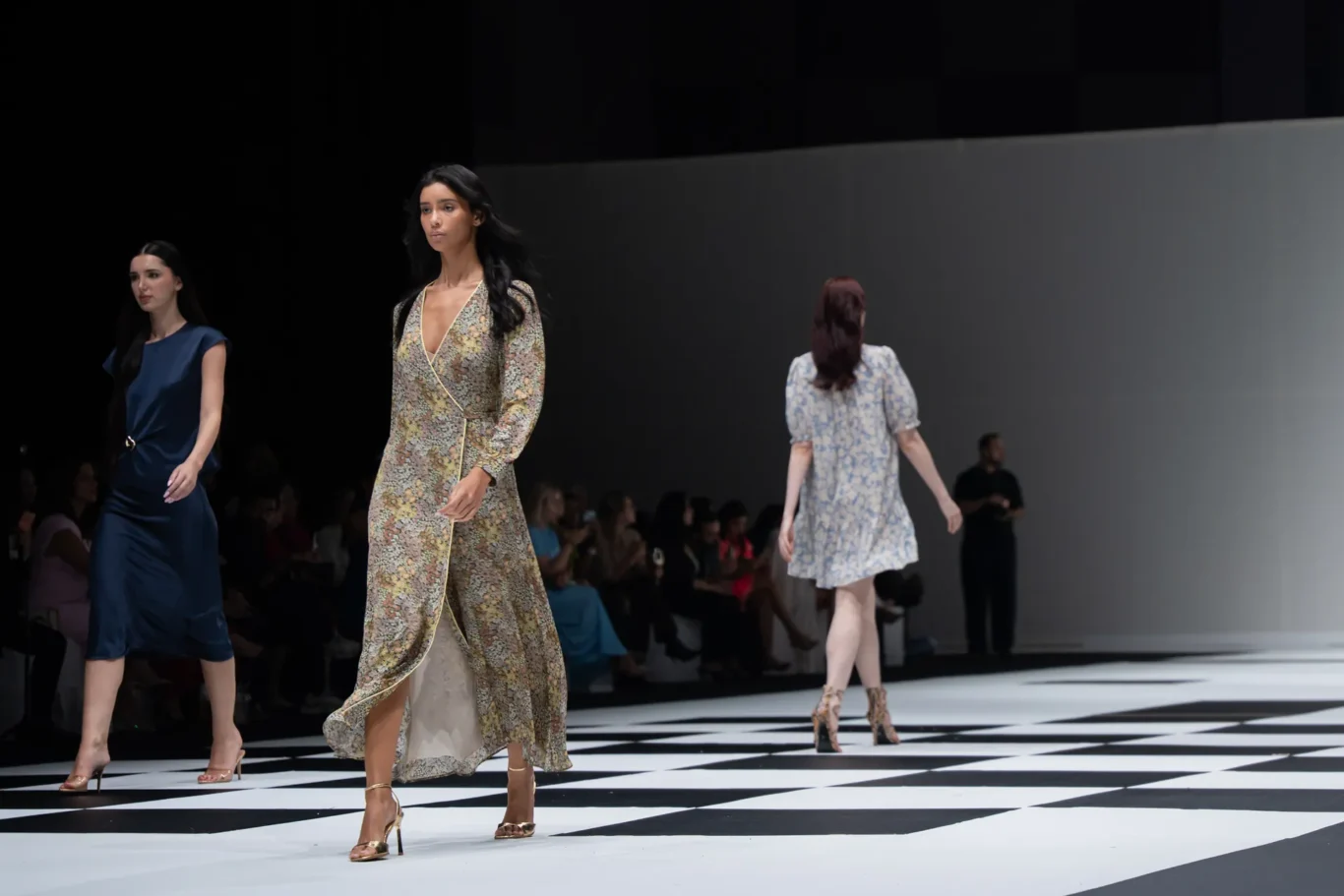 Models walk a fashion runway in floral, blue, and patterned dresses on a black-and-white checkered floor.