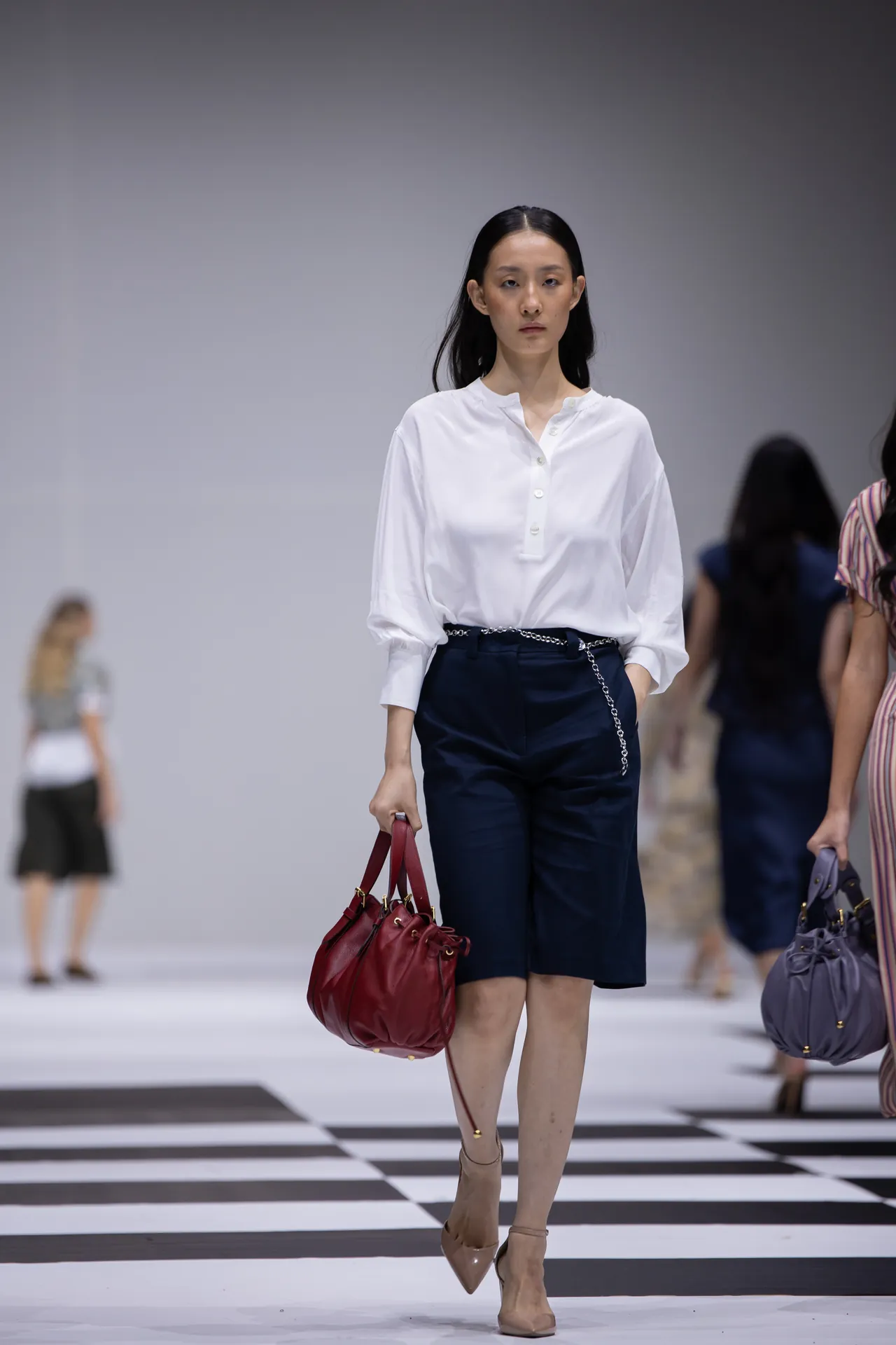Model walking a fashion runway in a white blouse, navy shorts, beige heels, and a red handbag.