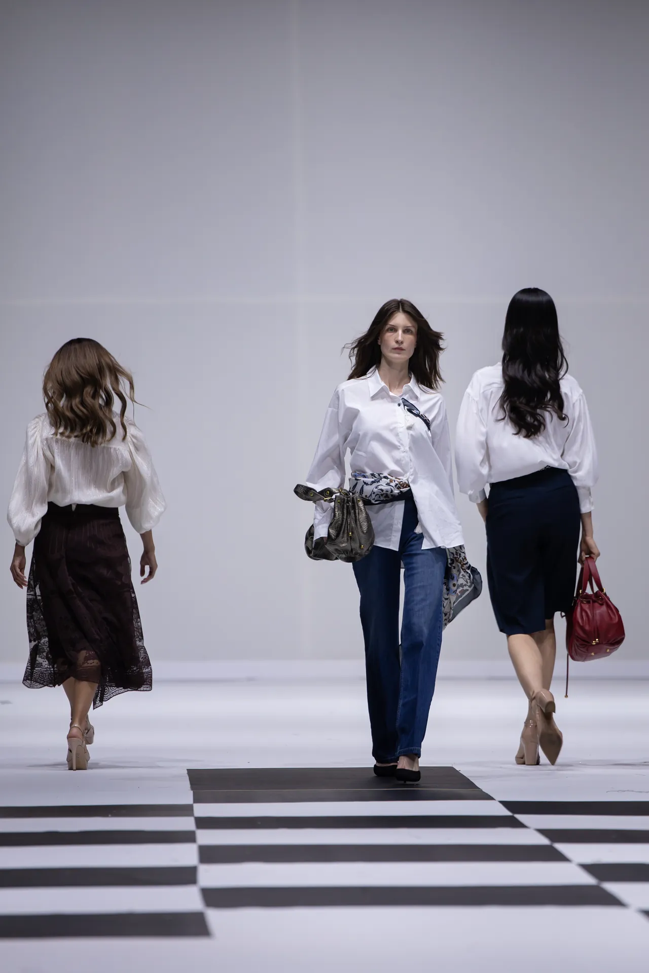 Three models walk a fashion runway on a black-and-white striped floor, wearing white blouses and carrying handbags.