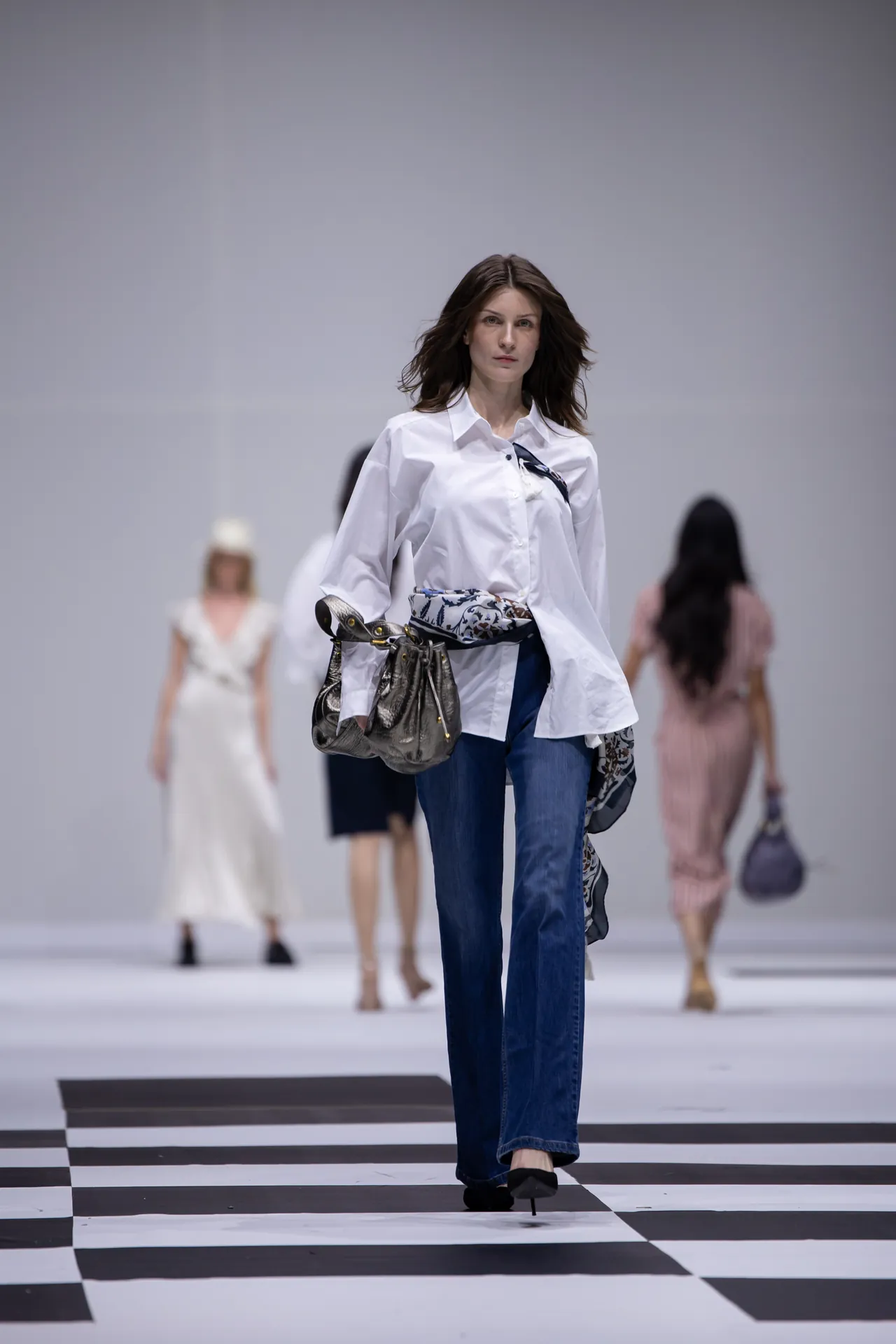 Female model walking a runway in a white button-down blouse and blue jeans, carrying a patterned scarf and metallic handbag.