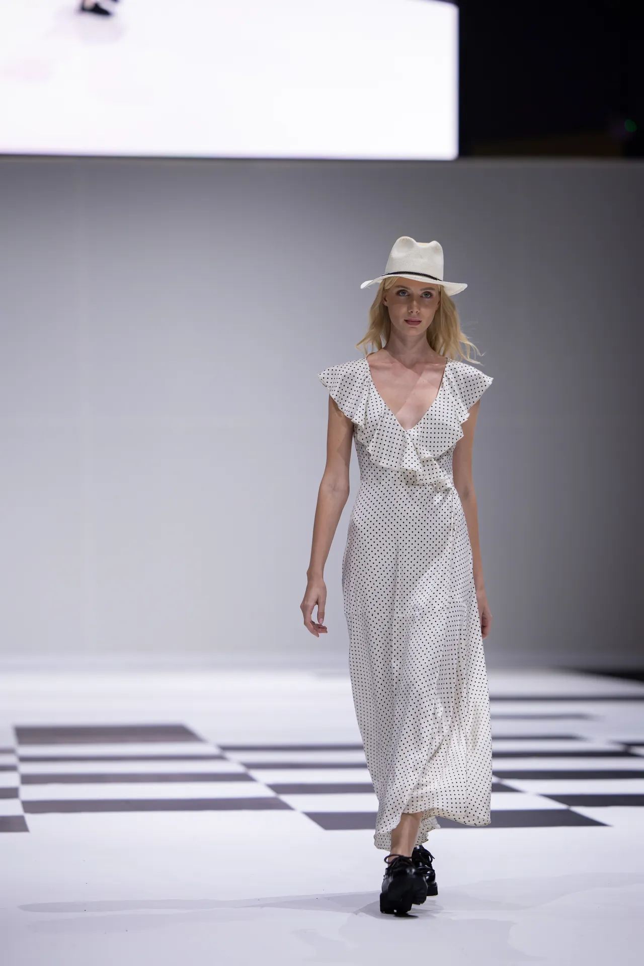 Runway model walking the catwalk in a white polka-dot dress with a white hat on a black-and-white checkered floor.