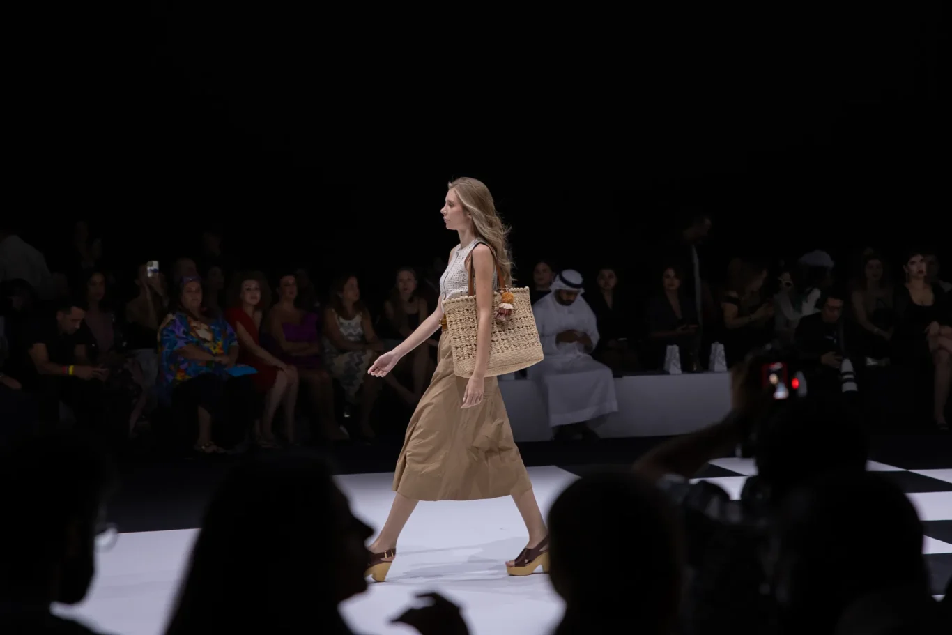 Model walks the runway wearing a white sleeveless top and tan skirt, carrying a large woven straw tote.