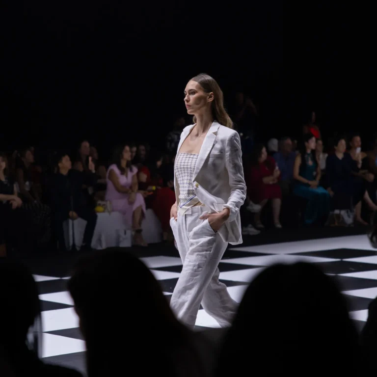 Model in a white blazer and pants walking a black-and-white checkered runway at a fashion show with an audience watching.",