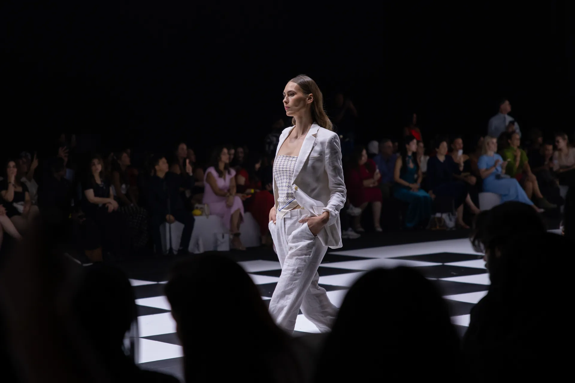 Model in a white blazer and pants walking a black-and-white checkered runway at a fashion show with an audience watching.",