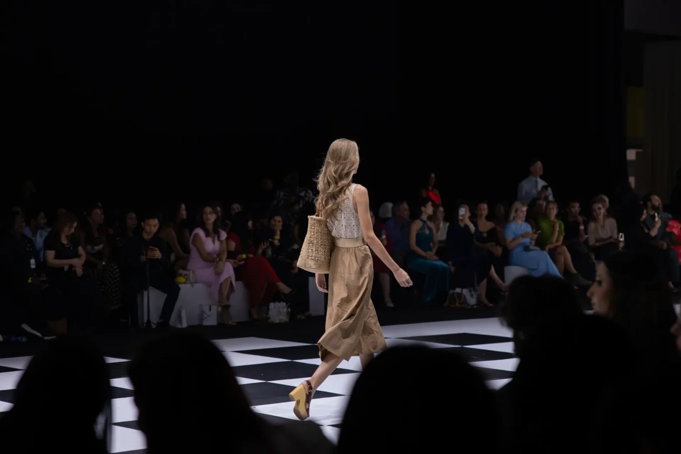 Model walks down a black-and-white checkered runway in a beige outfit, carrying a woven tote, as seated spectators watch.