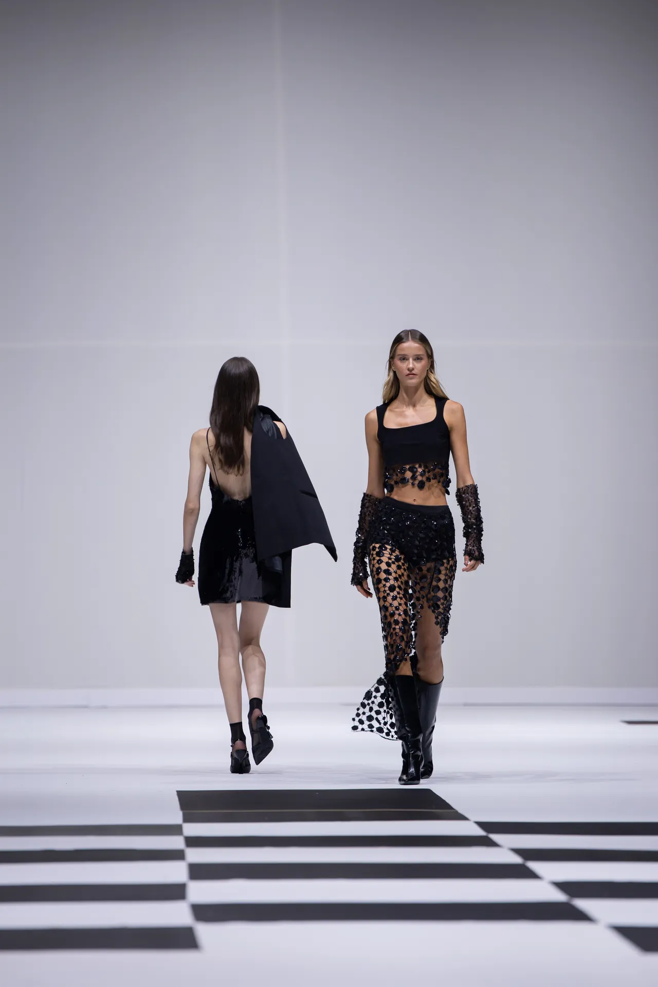 Two models walk a fashion runway in black outfits on a white stage with a bold black-and-white checkered floor pattern.