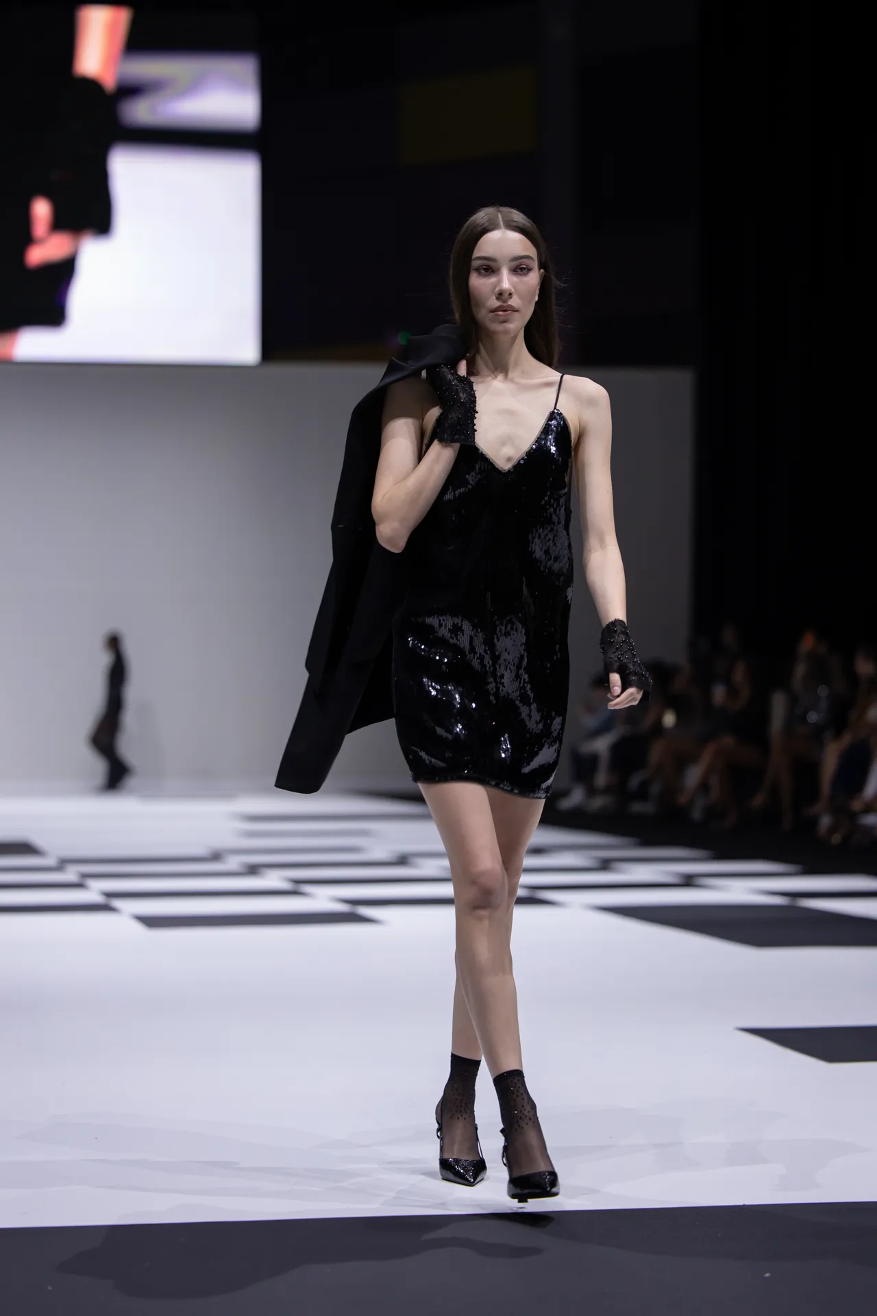 Model walks the runway in a black sequined slip dress, blazer draped over one shoulder, on a checkered catwalk nears the audience.
