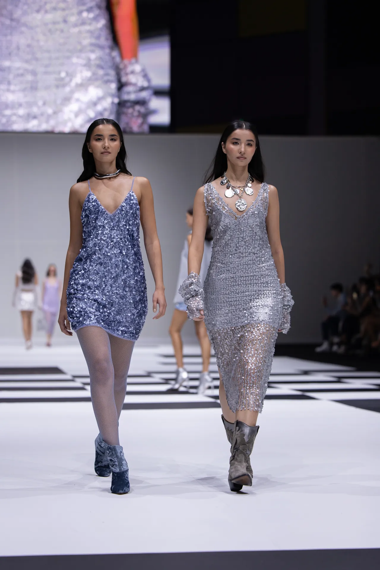 Two models walk a white runway in blue and silver sequin dresses during a fashion show (glittering outfits).