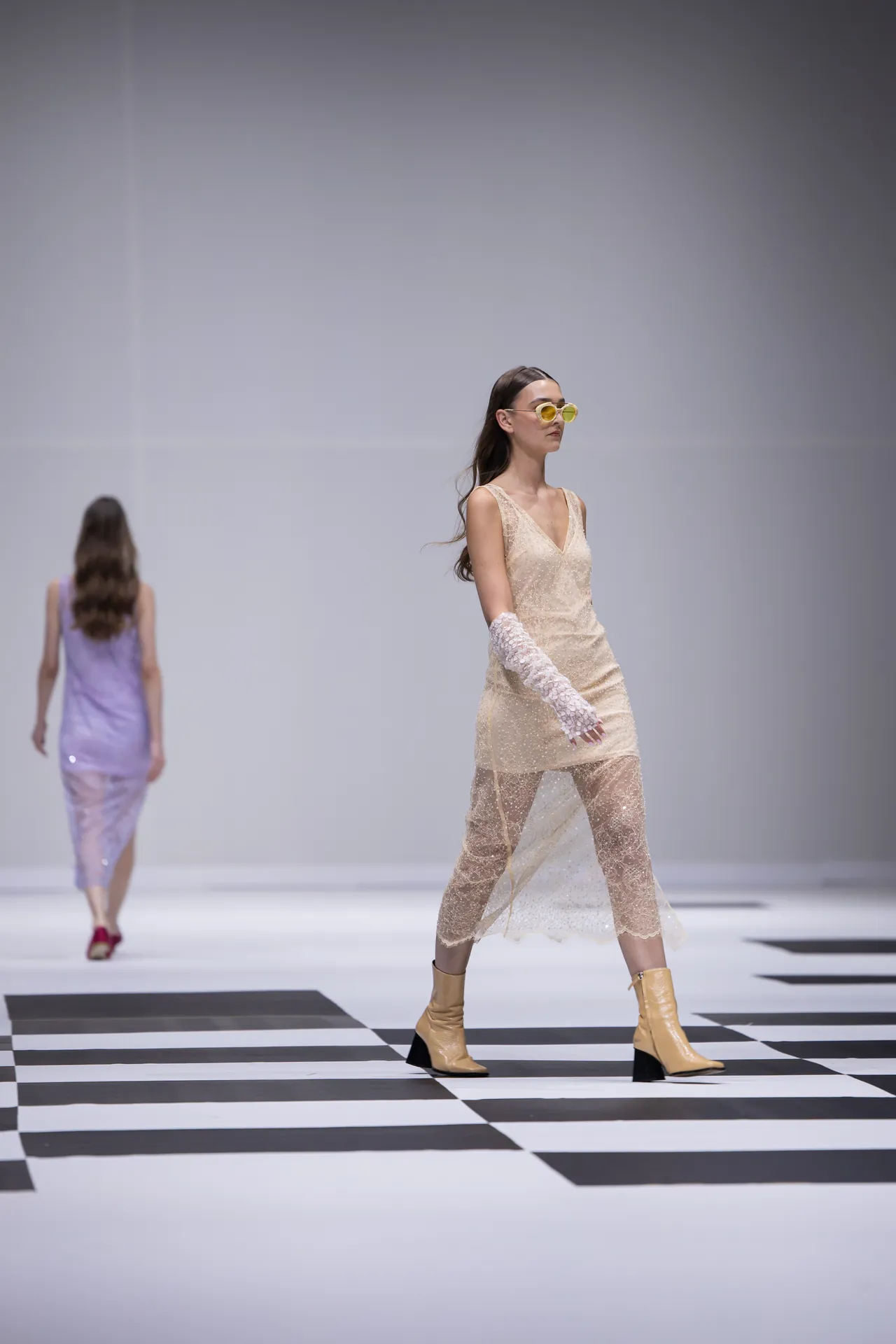 Model walks a runway in a beige lace midi dress with sheer overlay, yellow sunglasses, and lace gloves, beige boots on a black-and-white checkered floor with a blurred lavender-dressed model in the background