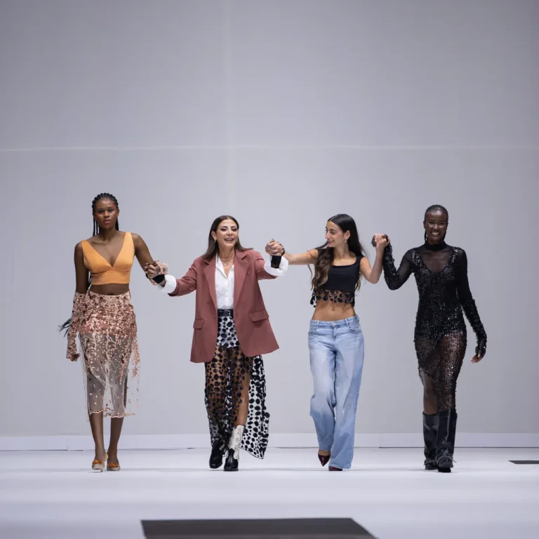 Four diverse models walk hand-in-hand on a fashion runway with a black-and-white striped floor.