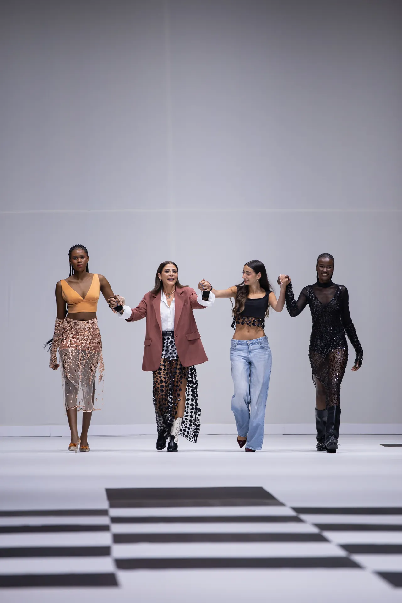 Four diverse models walk hand-in-hand on a fashion runway with a black-and-white striped floor.