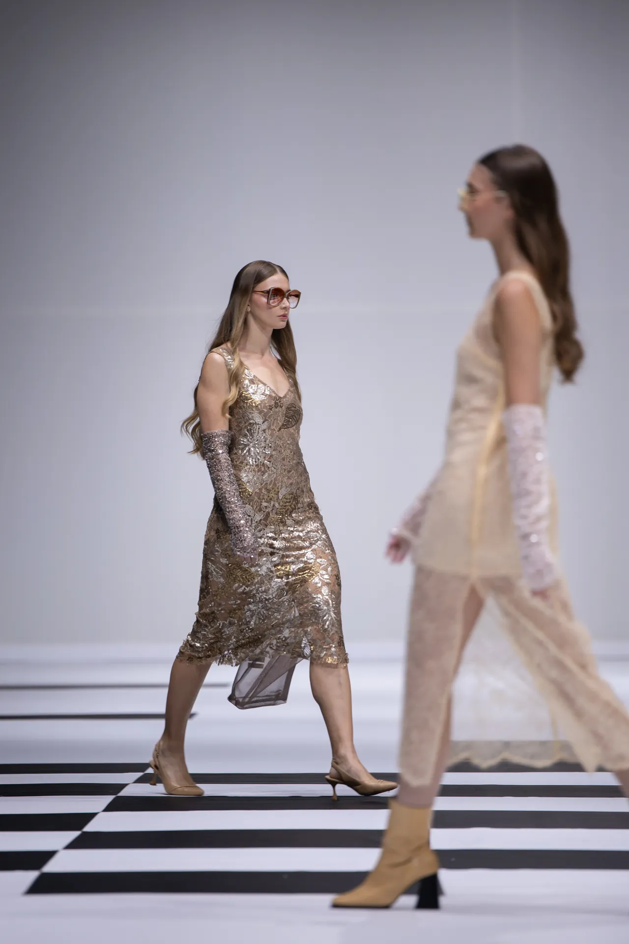 Model walks a fashion runway in a gold sequin dress with matching glittery arm sleeves, wearing sunglasses on a black-and-white checkered floor.