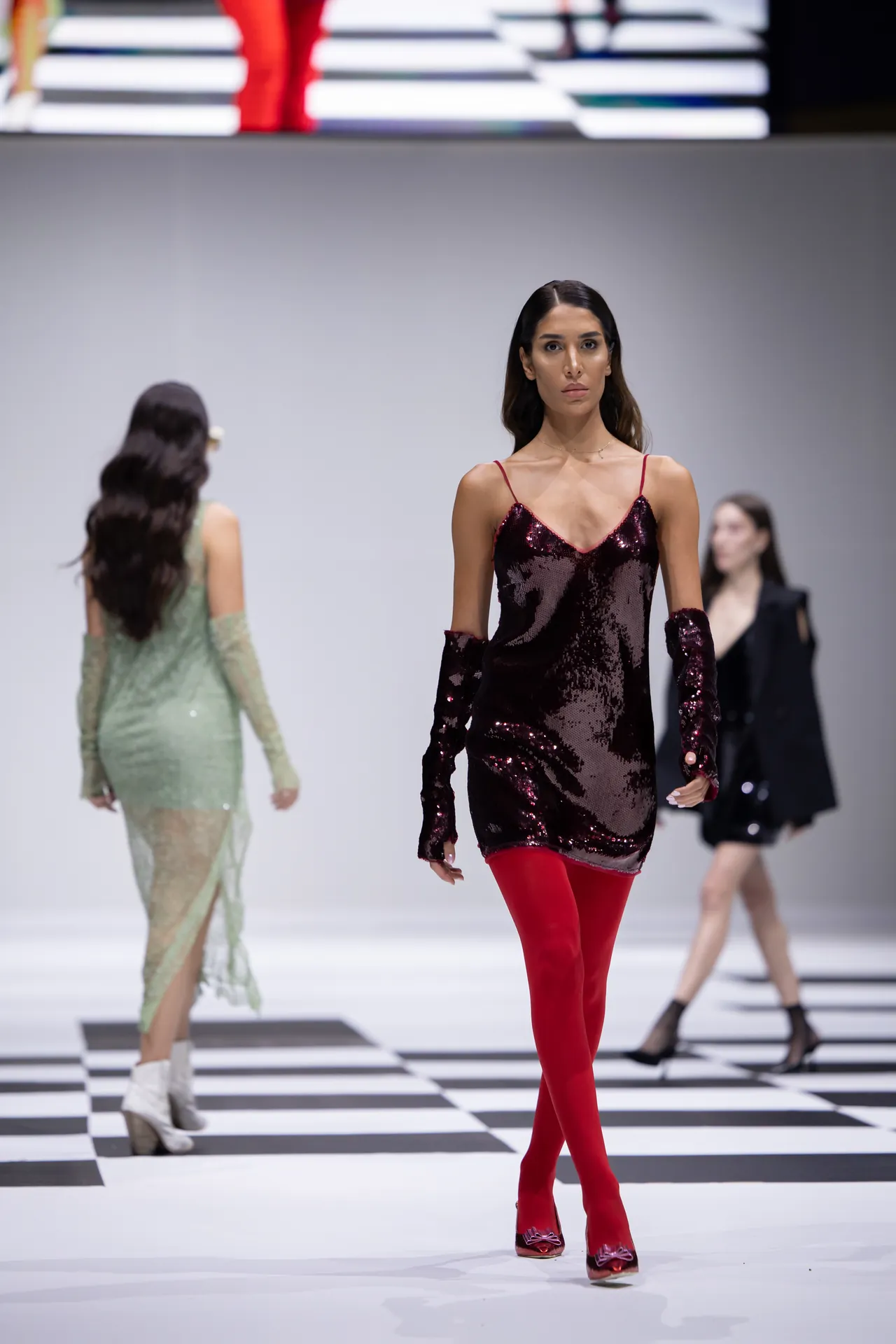 Model walks the runway in a burgundy sequin mini dress with red tights and matching shoes, foreground focus on outfit.