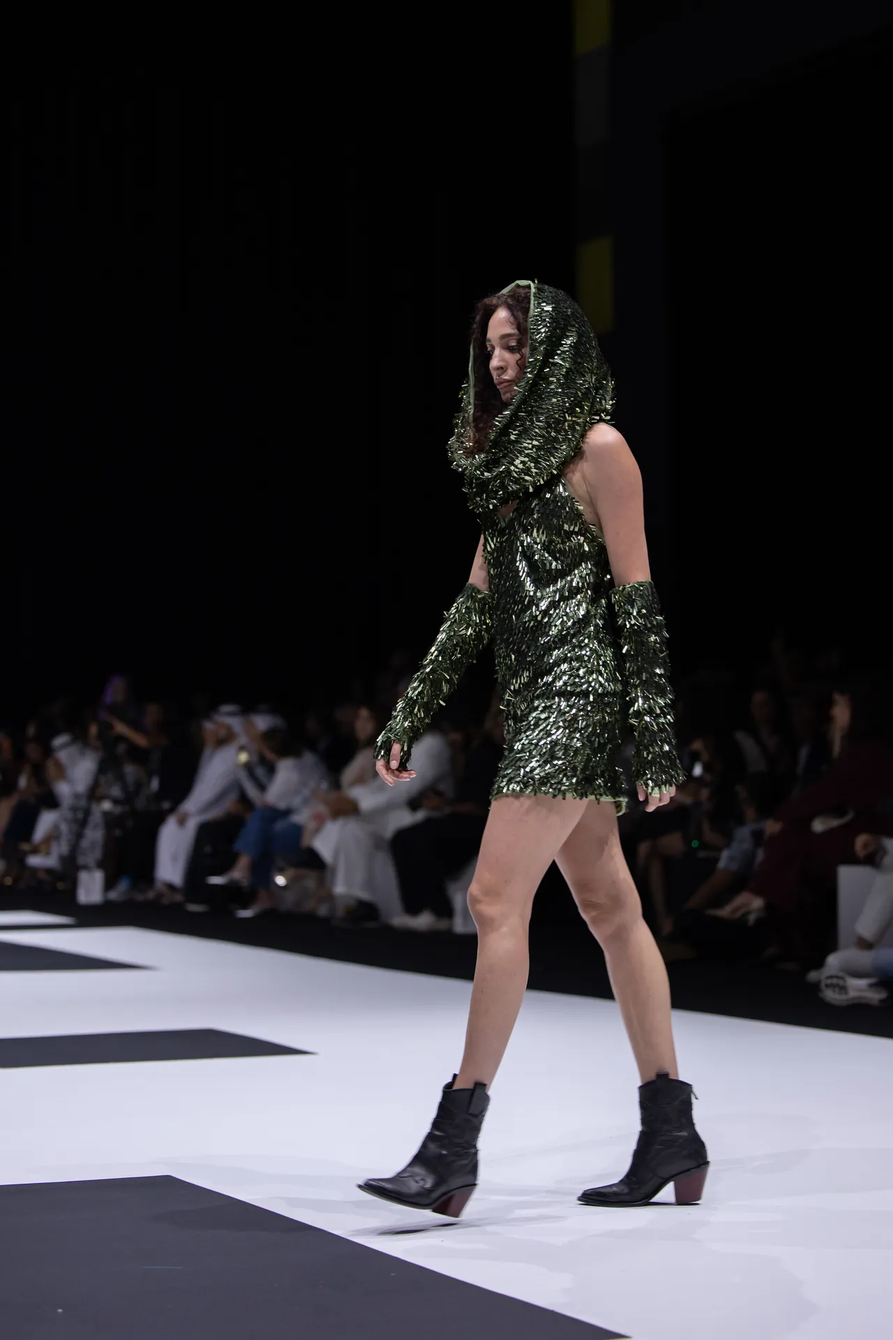 Model walking a fashion runway in a green sequin hooded dress with gloves and black ankle boots, audience in background.