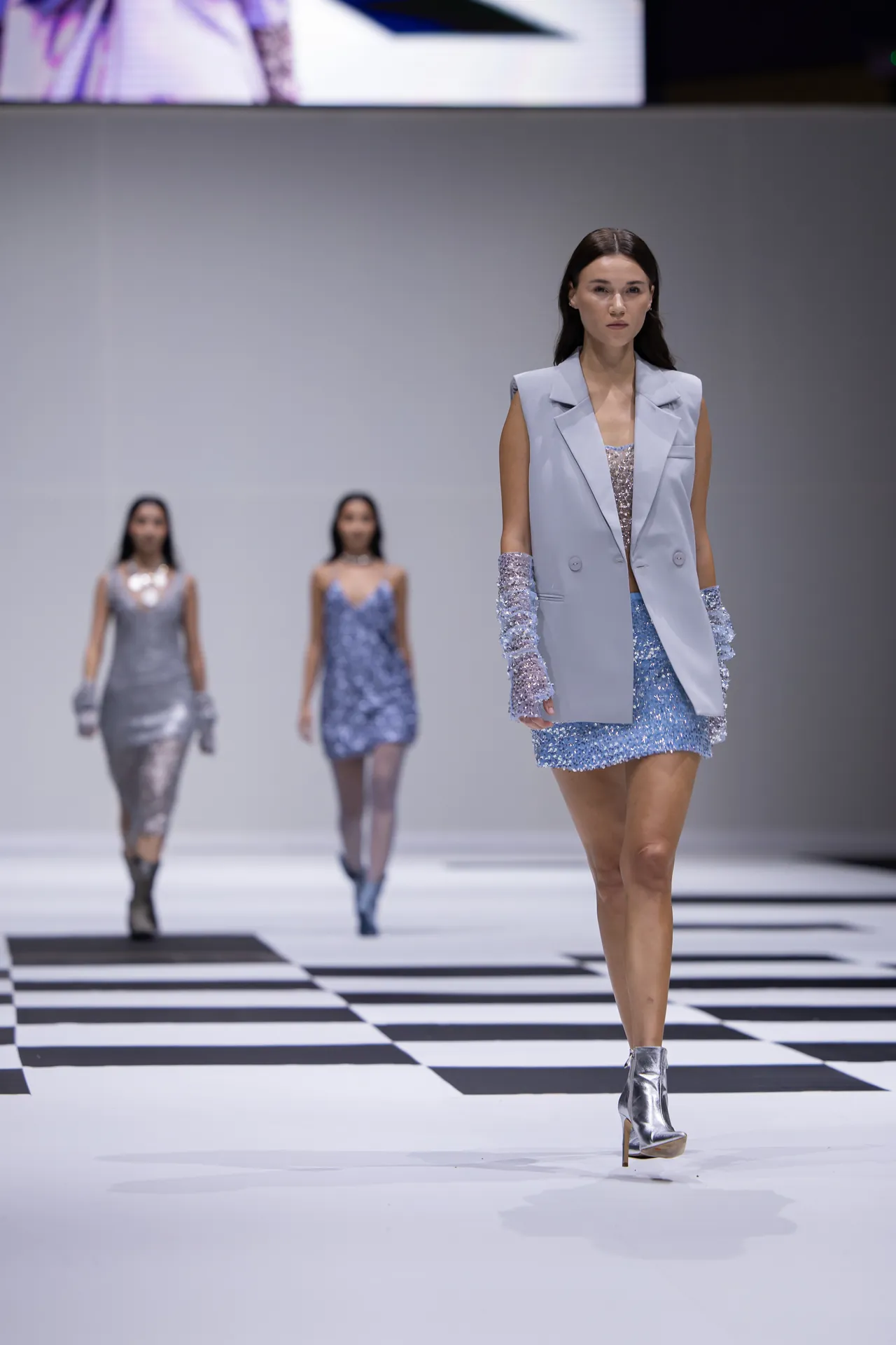 Model walks the runway in a pale blue sleeveless blazer over a blue sequin mini dress, silver ankle boots on a black-and-white striped catwalk.