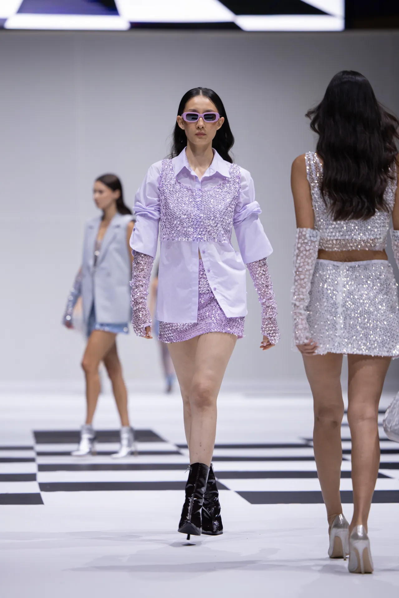 Model walking a fashion runway in a lavender sequin outfit with matching sleeves and lavender sunglasses against a black-and-white checkered runway newsprint backdrop.
