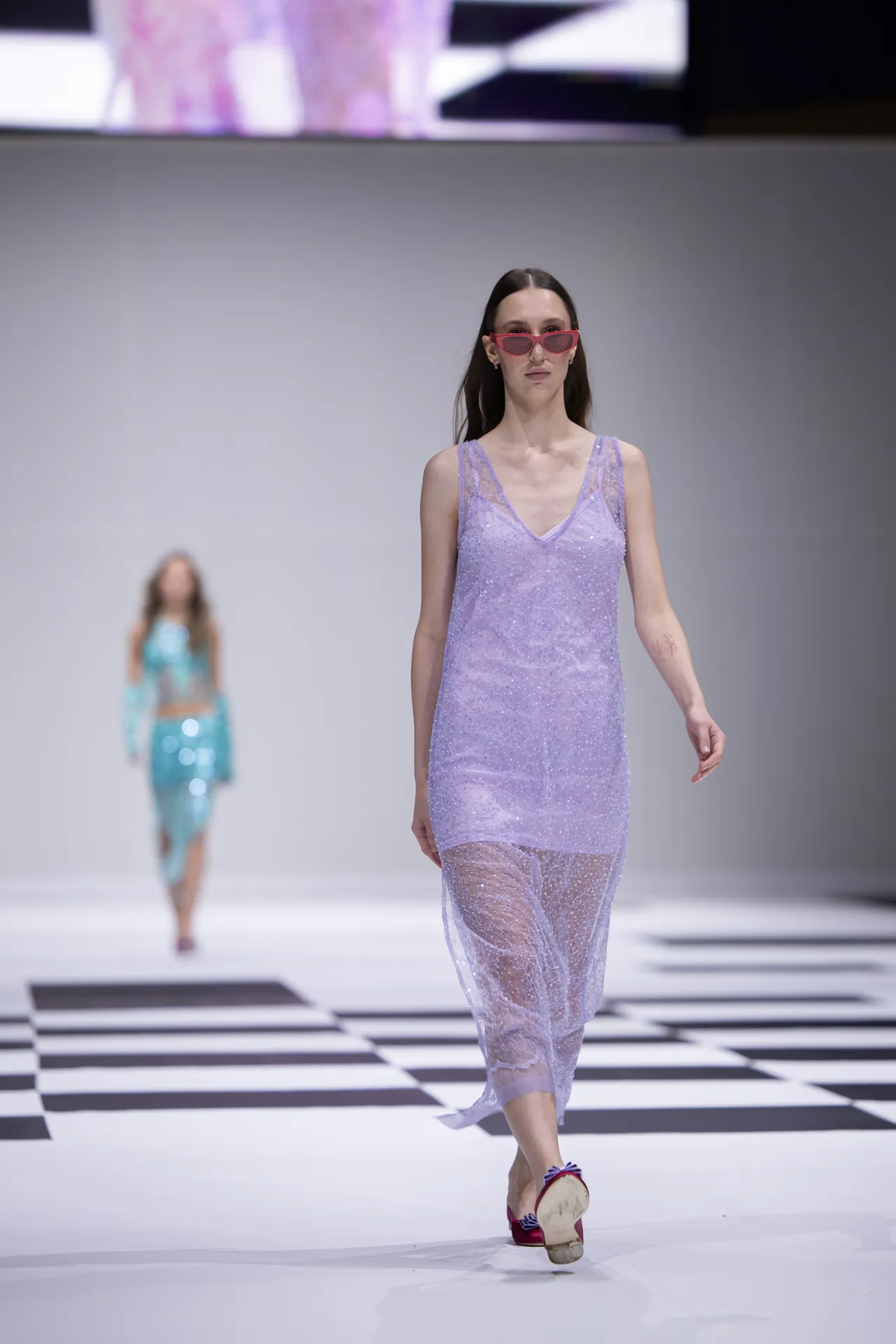 Model on a fashion runway wears a lavender sheer, beaded dress and pink sunglasses; black-and-white checkered runway beneath.