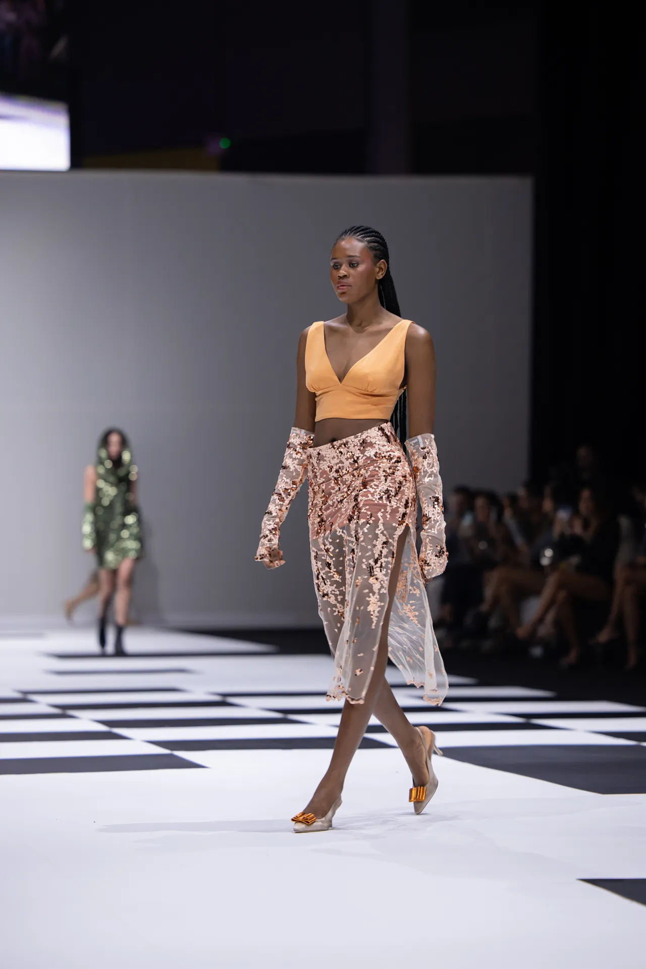 Model walks a fashion show wearing a peach crop top and sheer floral skirt with gloves, on a black-and-white checkerboard runway.