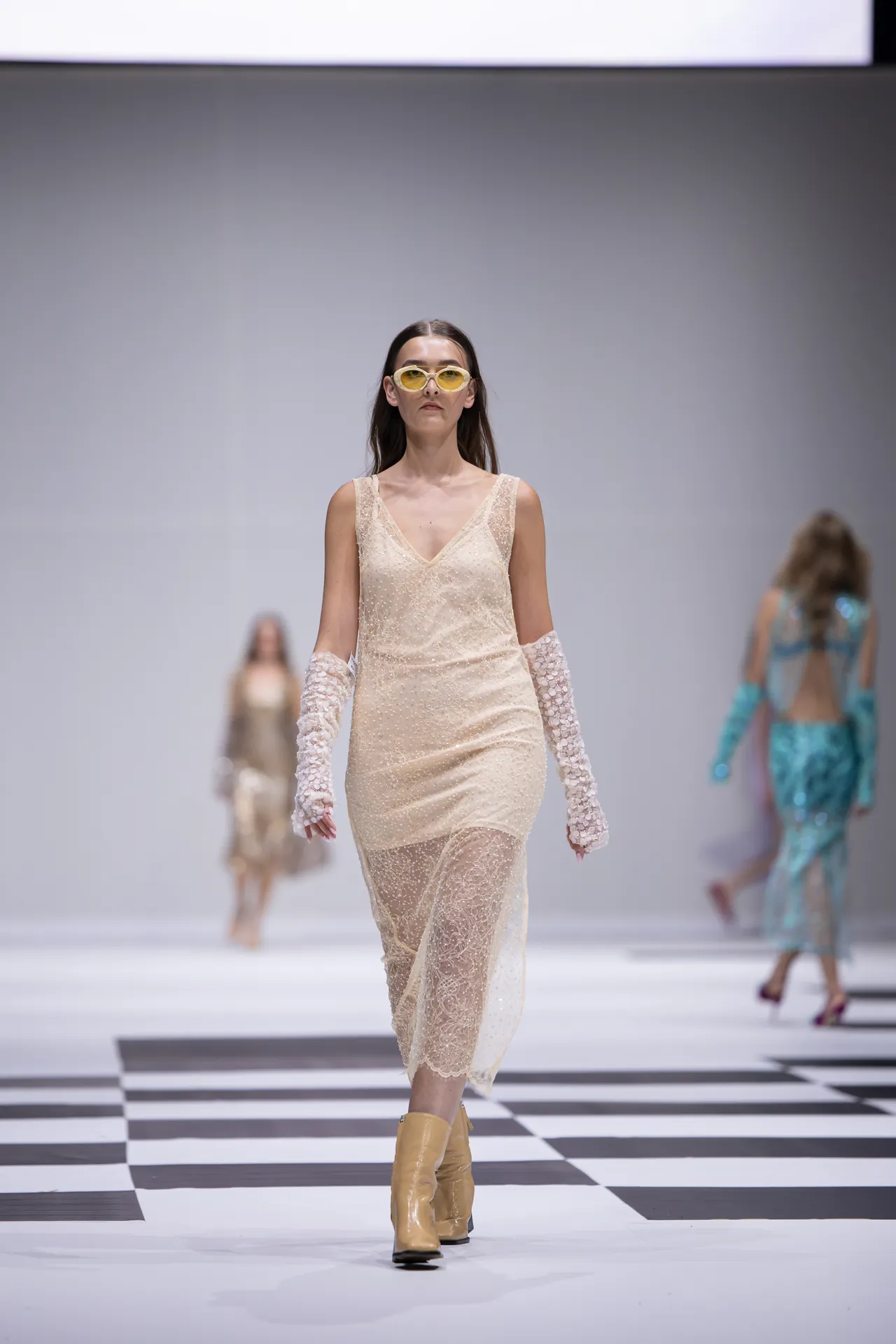 Model walks the runway in a cream lace slip dress with matching lace gloves and yellow sunglasses on a black-and-white checkered catwalk.