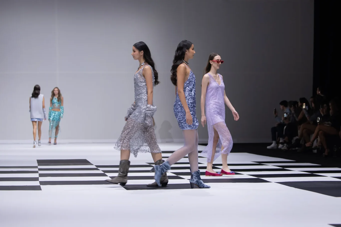 Models walk a runway in sparkling dresses on a black-and-white checkered floor, audience watching on the right.