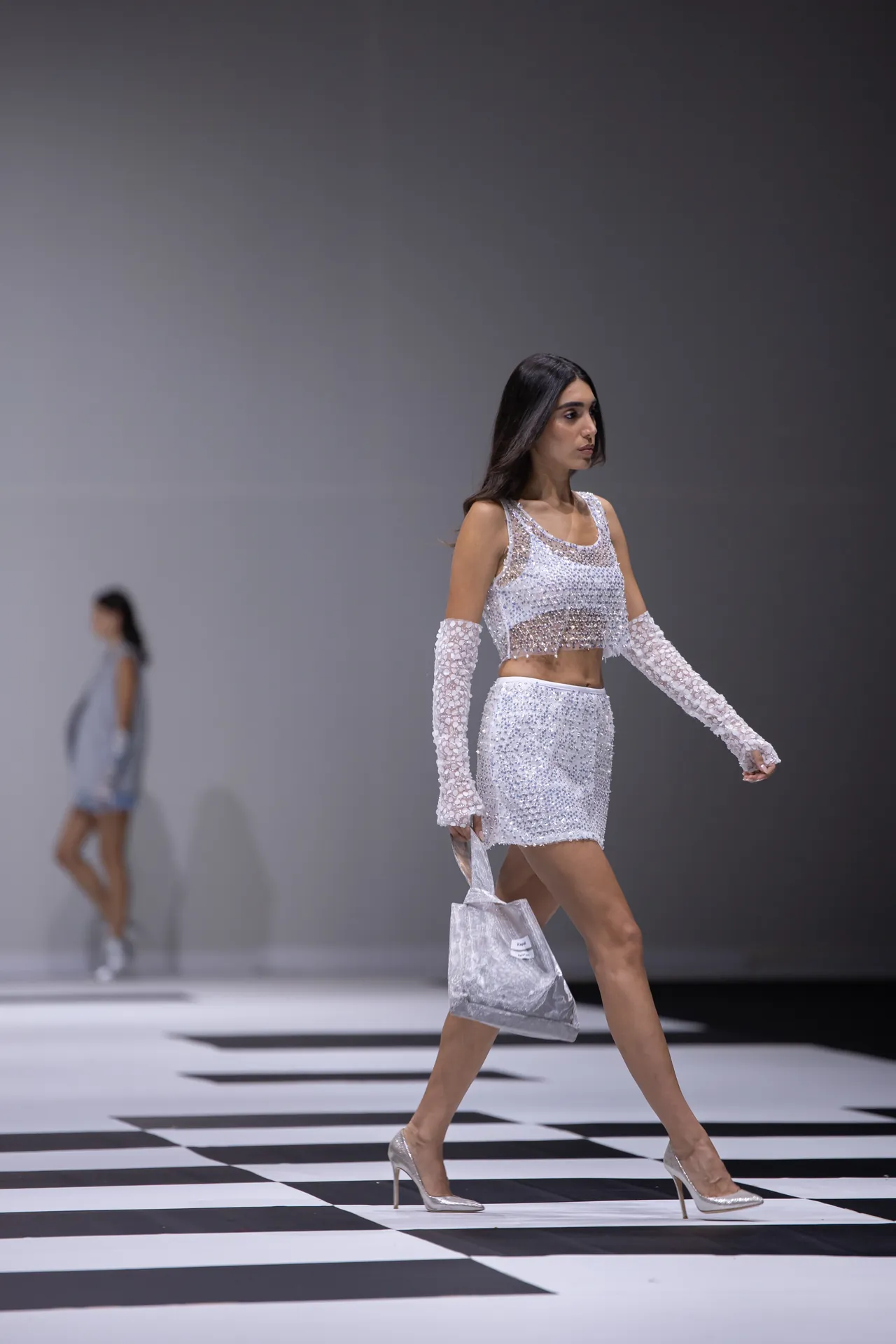 Model walking a fashion runway in a silver-sequined crop top and skirt with matching long gloves, carrying a metallic tote.