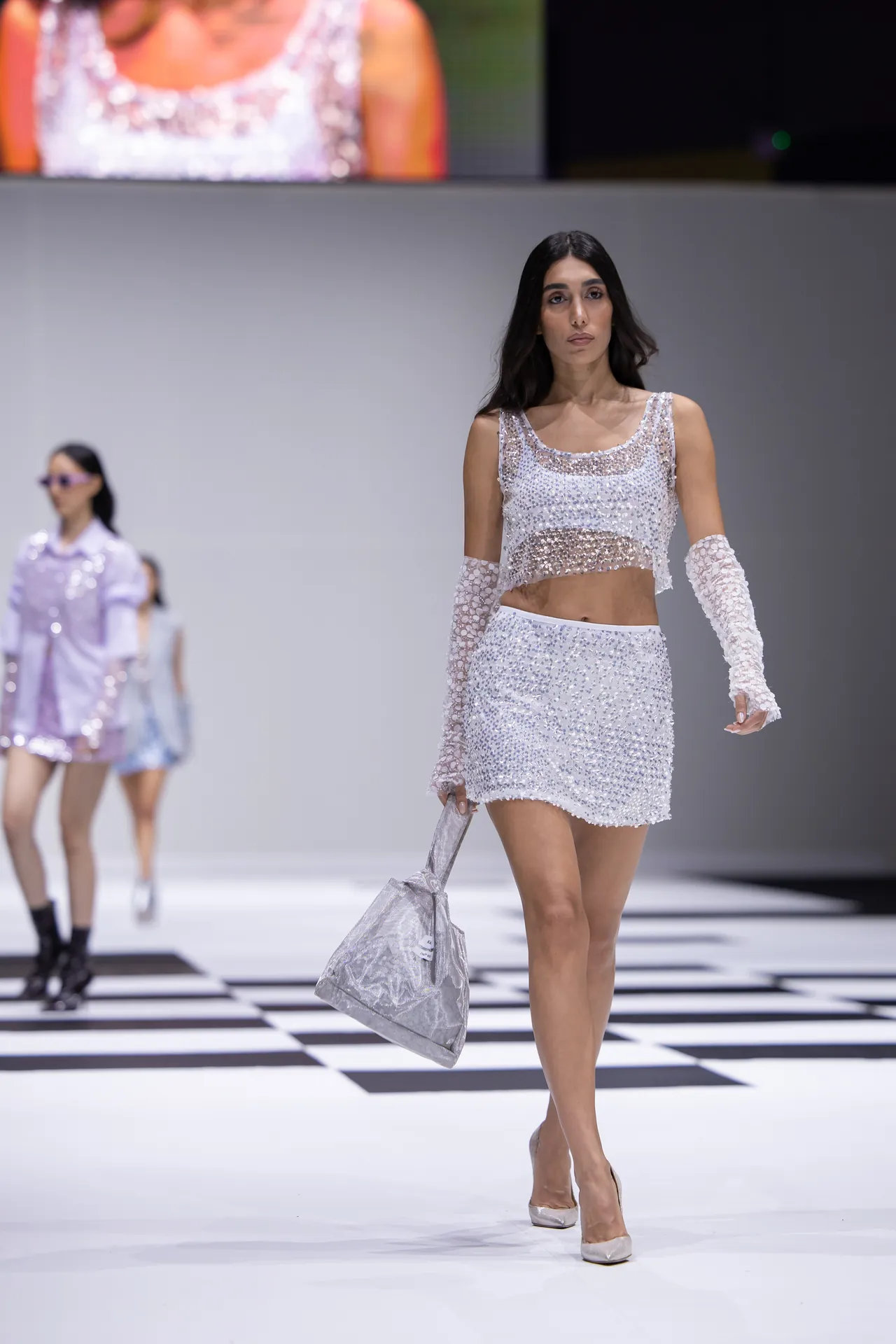 Model walking a silver sequin crop top and mini skirt on a checkered runway, carrying a metallic bag.