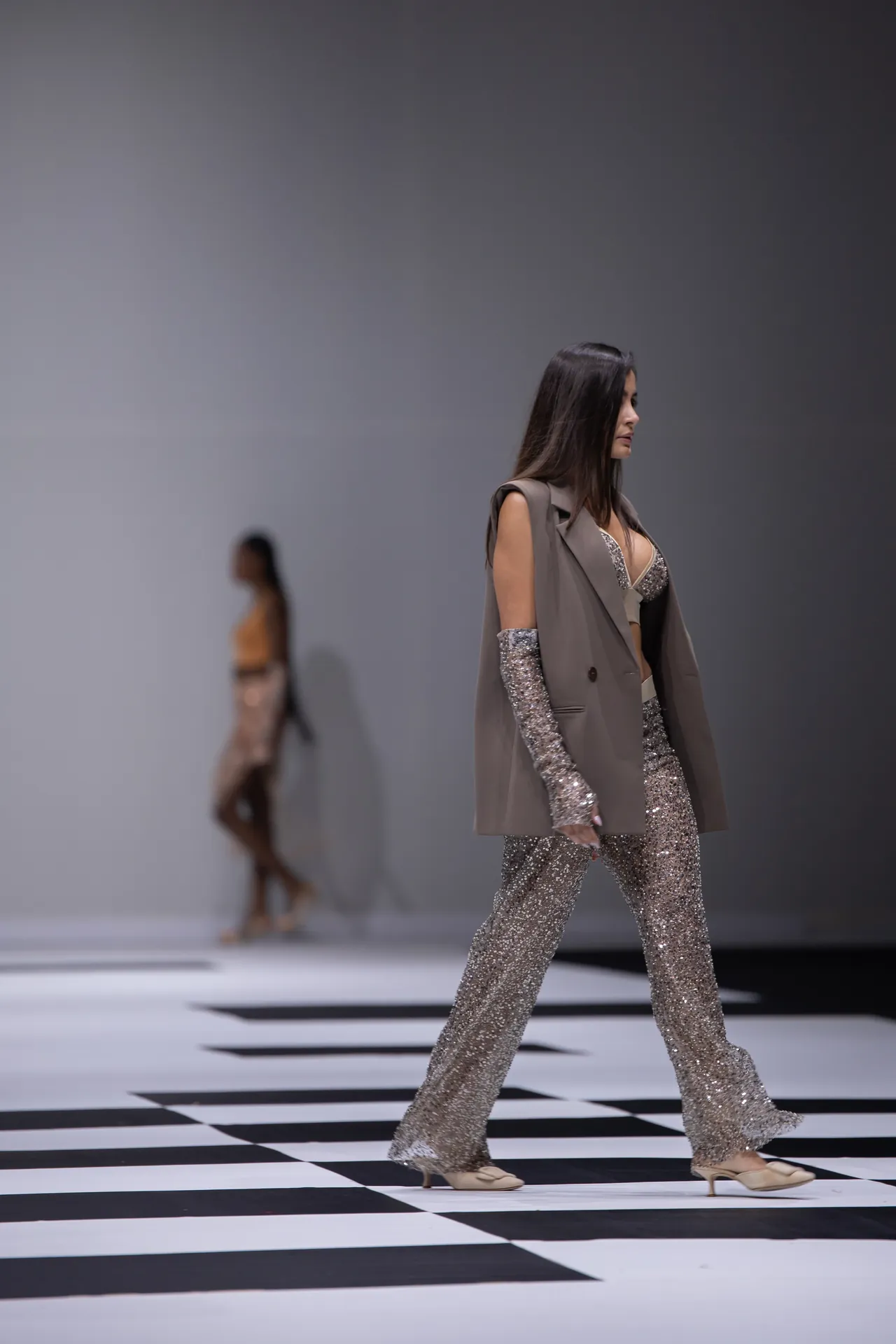 Model walking a fashion runway in a silver glitter jumpsuit and beige blazer, on a black-and-white checkered floor.