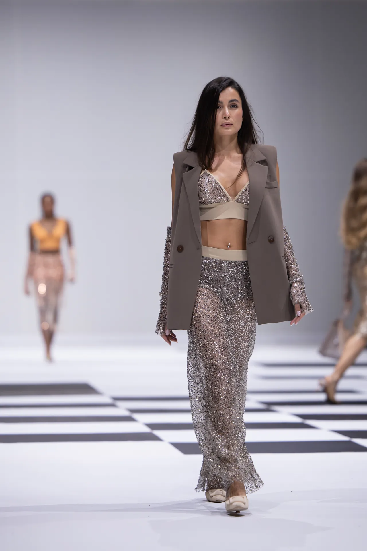 Model walking a fashion runway in a glittery silver two-piece outfit with a taupe blazer, on a black-and-white striped catwalk.