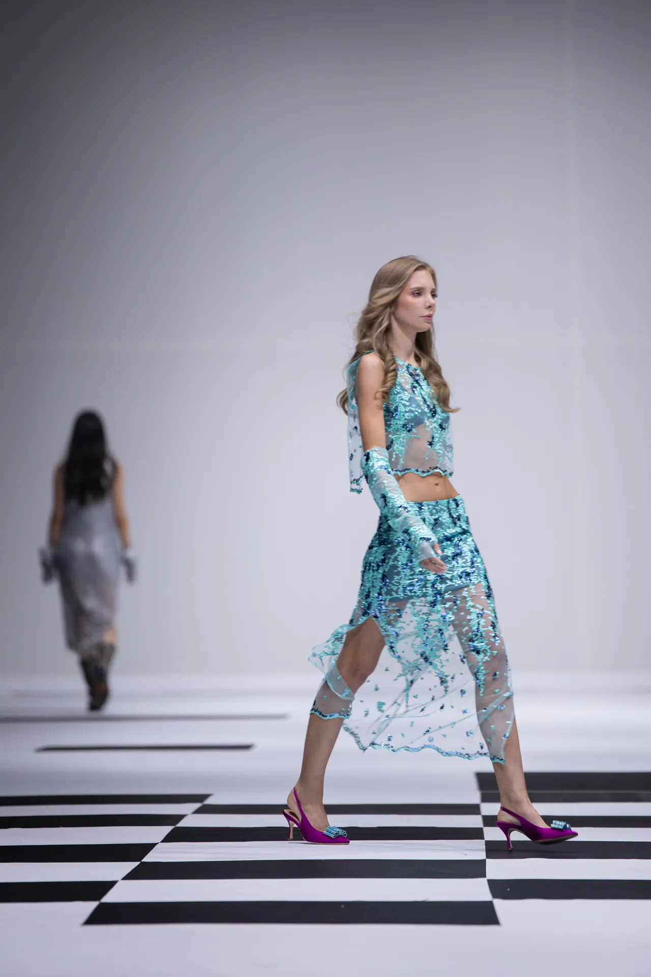 Model walking the runway in a turquoise sequin mesh outfit with gloves on a black-and-white checkerboard floor, with a blurred second model in the background.