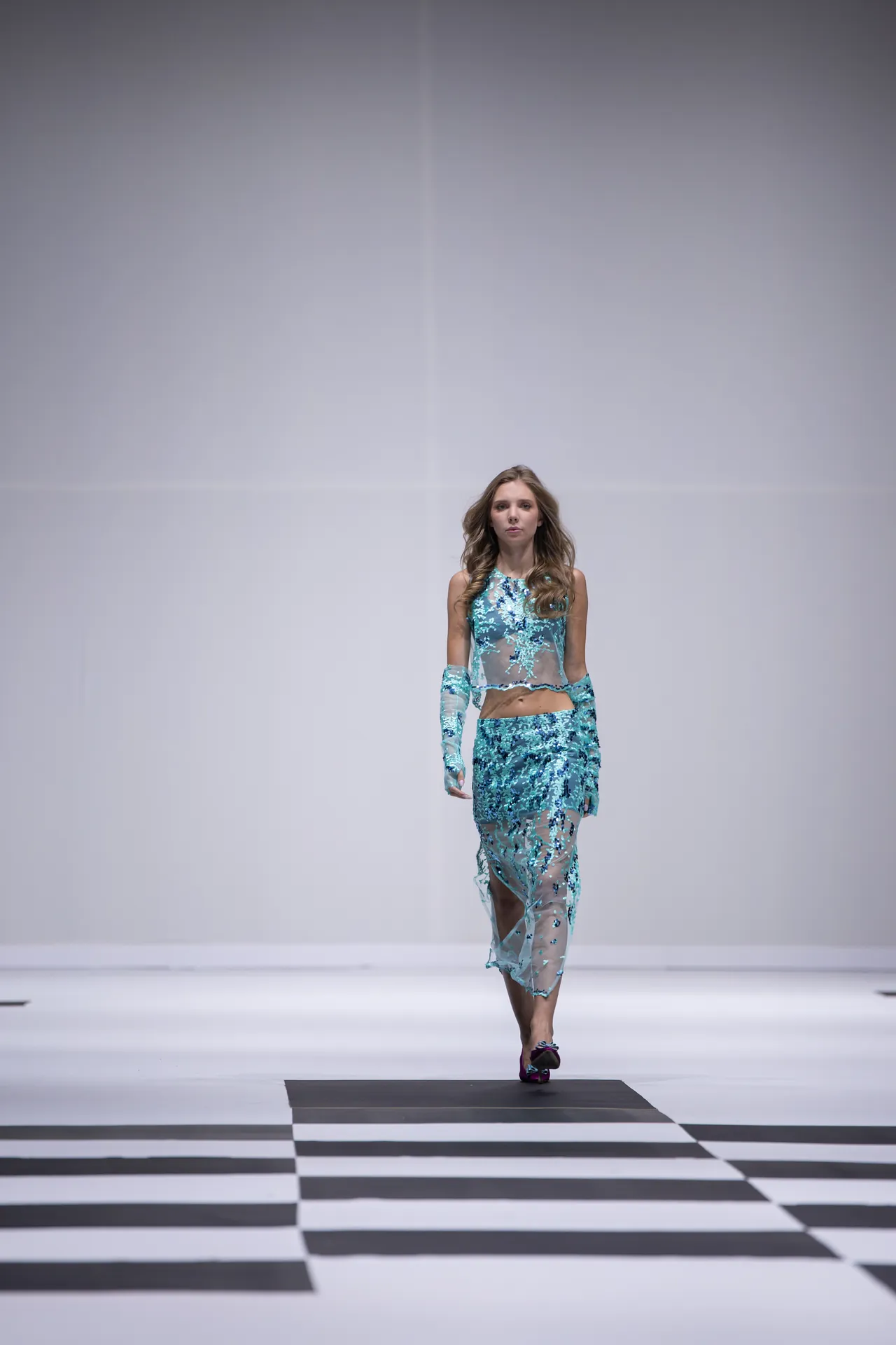 Model walking a runway in a turquoise floral sheer dress with long sleeves, on a black-and-white checkered catwalk.