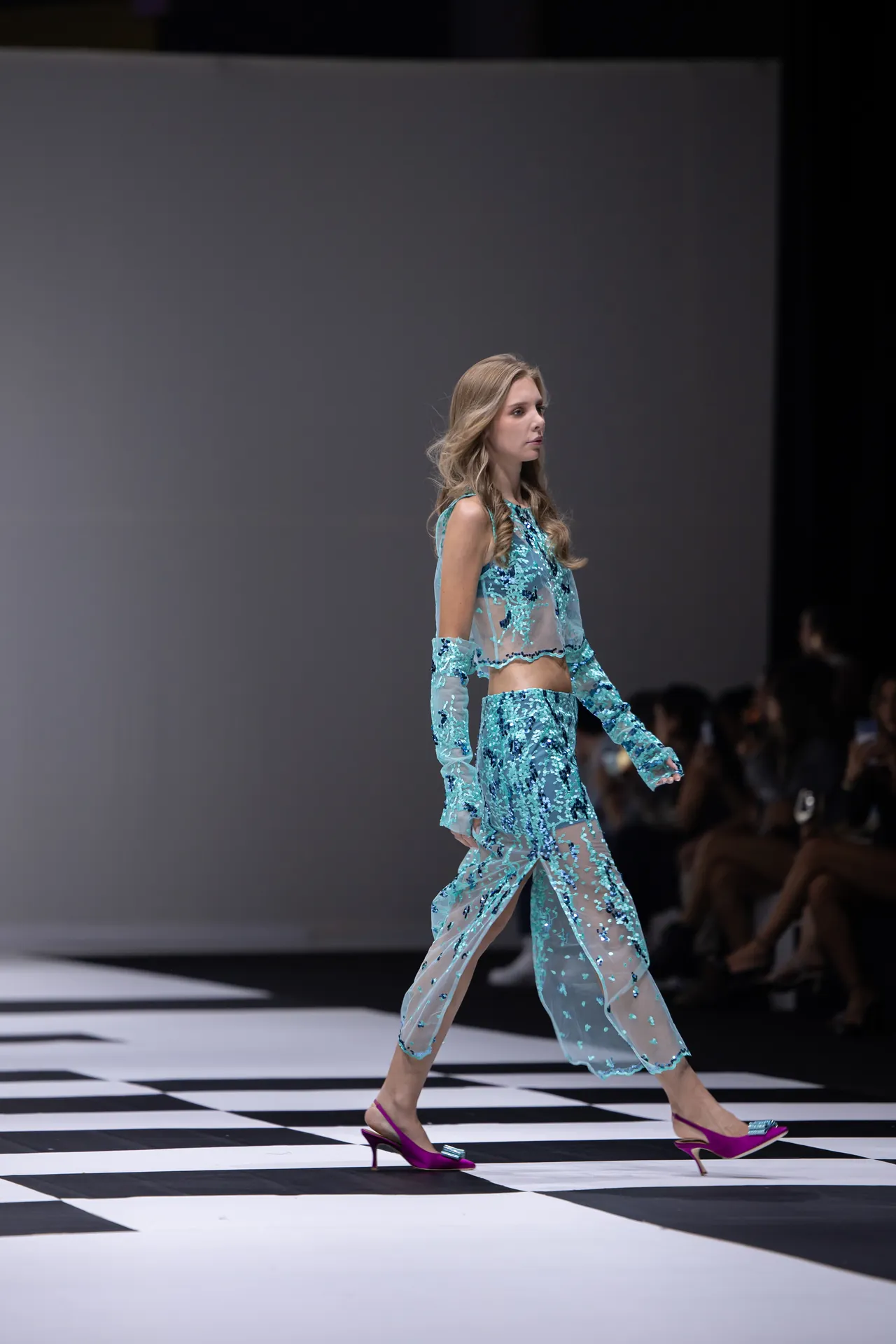 Female model walks the runway in a turquoise sheer sequined two-piece outfit, mid-stride on a black-and-white checkered runway floor.