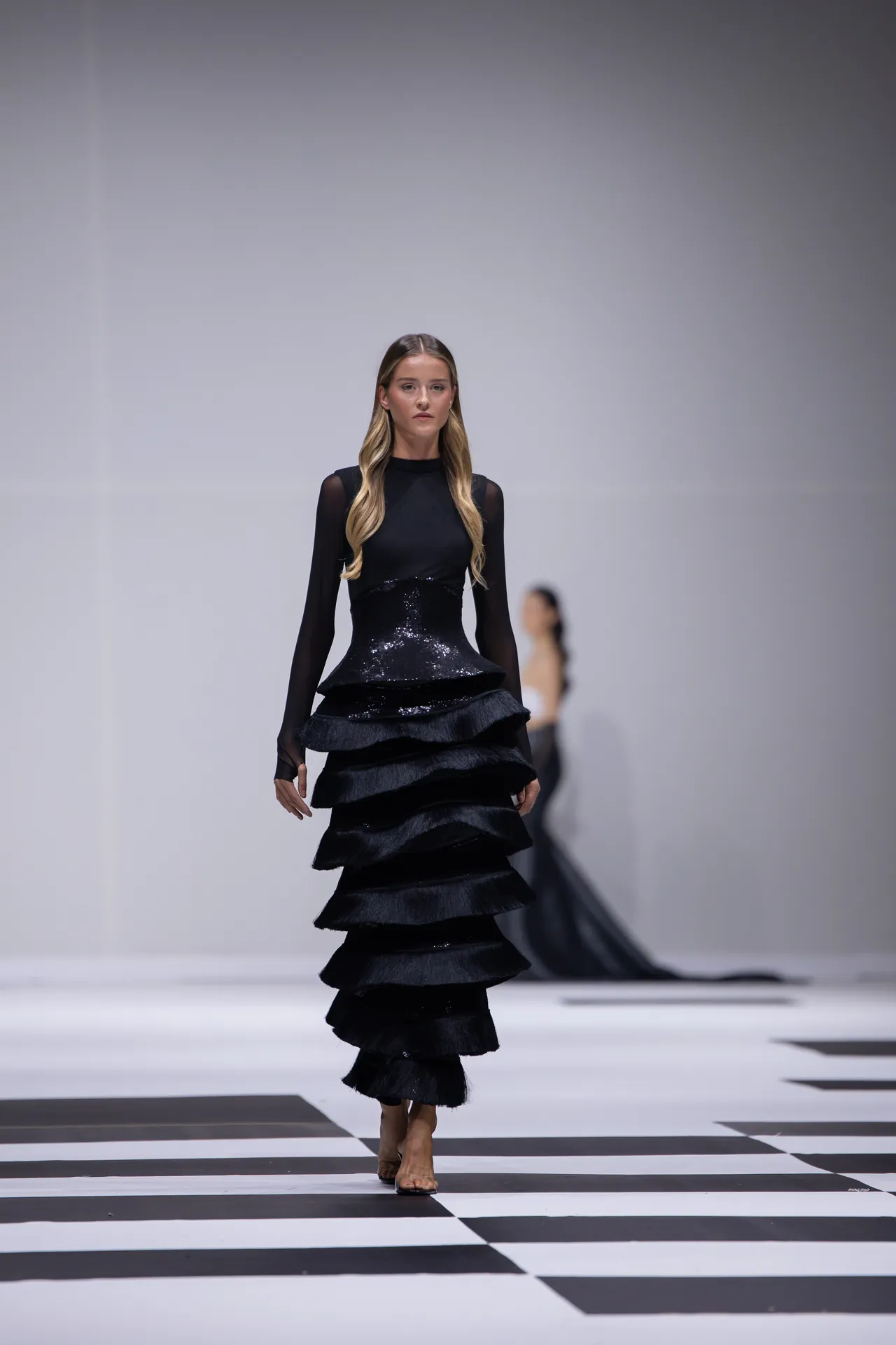 Model walks a black, multi-tiered sequin gown on a black-and-white checkerboard runway image.