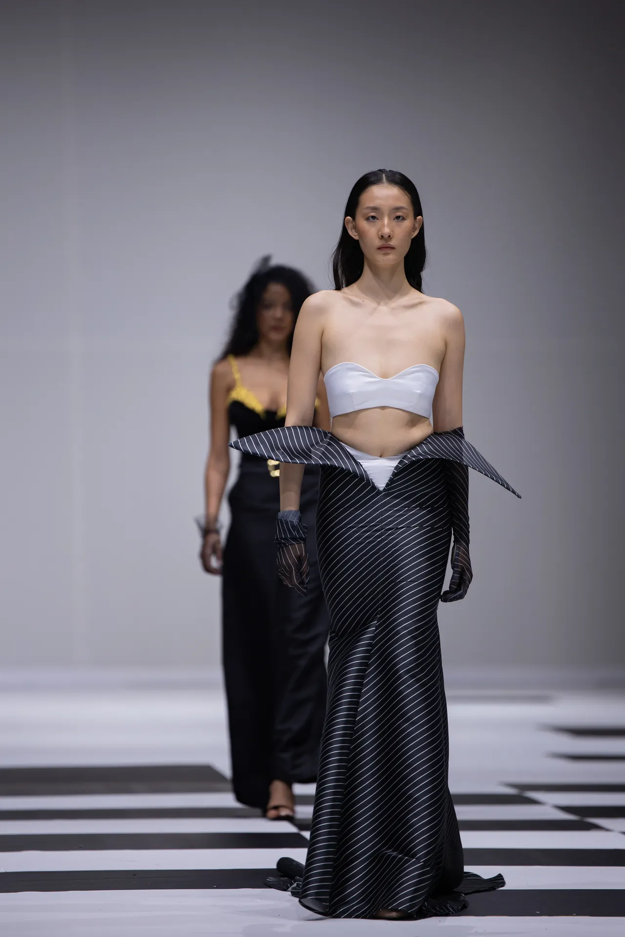 Model walks a fashion runway in a white strapless bustier top and a black pinstriped skirt with a sculptural, folded waistband.