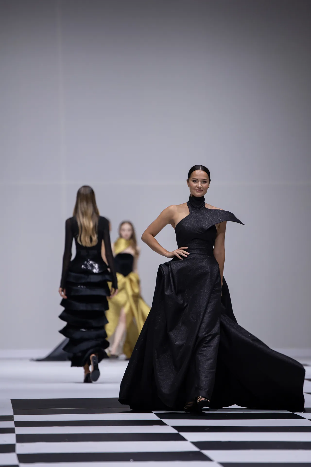 Model in a shimmering black gown walks the runway, with a black-and-white checkered floor and other models in the background.