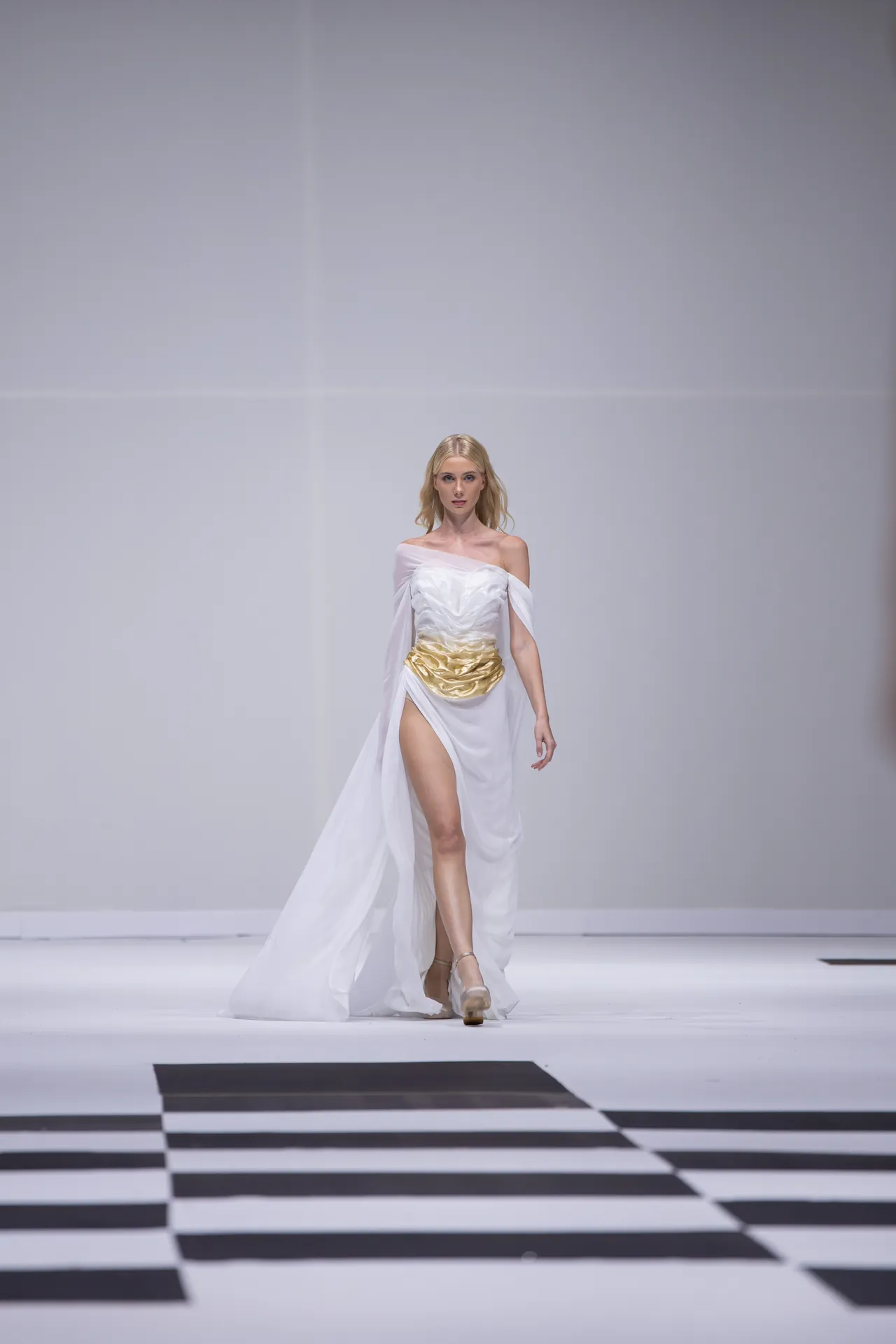 Model walks a fashion runway in a white off-shoulder gown with a gold waist belt, on a black-and-white checkered catwalk.