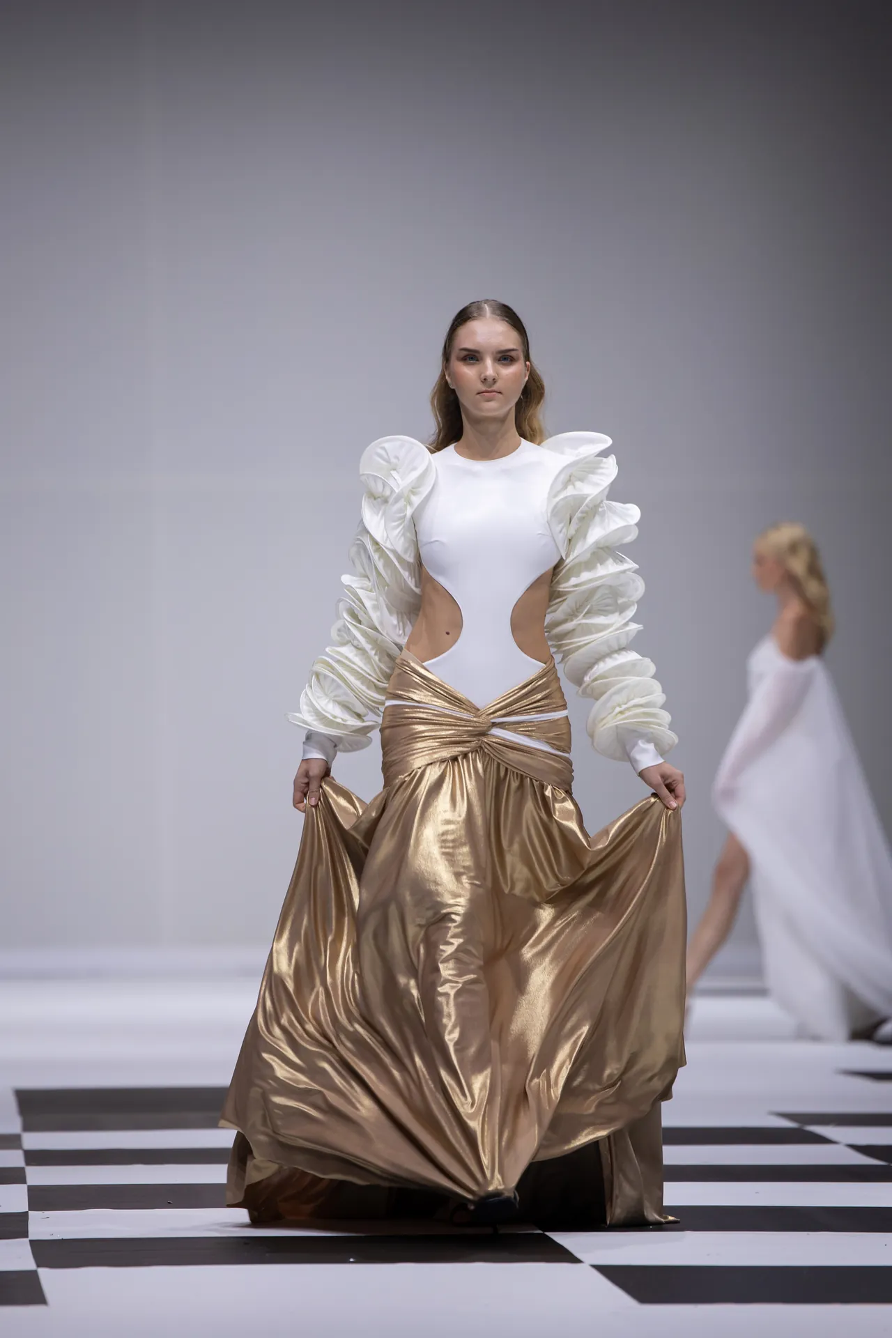 Model on a runway wearing a white bodysuit with oversized sculptural puff sleeves and a metallic gold draped skirt.