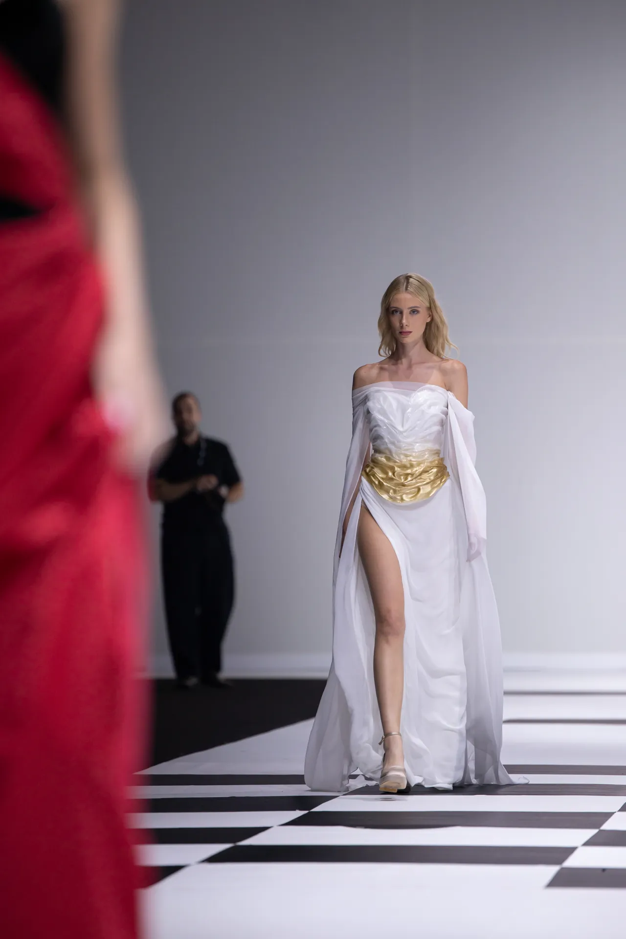 Model walking a fashion runway in a white off-shoulder gown with a gold waist sash on a black-and-white striped catwalk, foreground blurred red garment present.