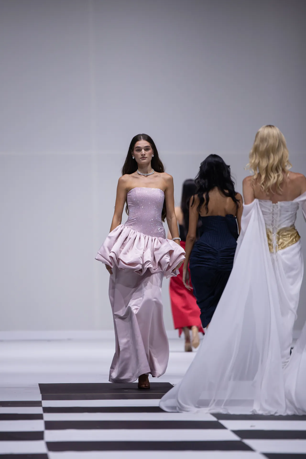 Model walks a fashion runway in a lavender strapless gown with a large ruffled skirt on a black-and-white checkered floor.