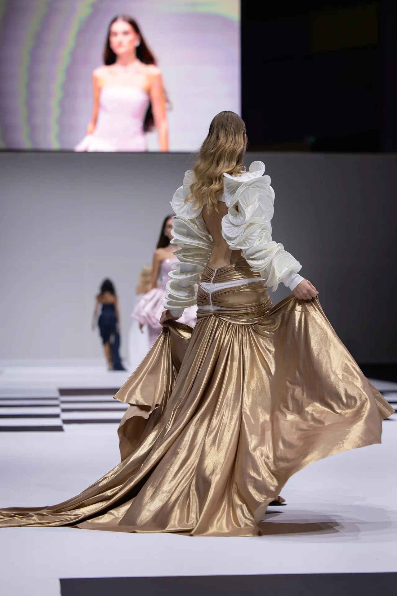 Model on a runway wearing a gold satin gown with dramatic white ruffle sleeves, back view as other models walk behind.