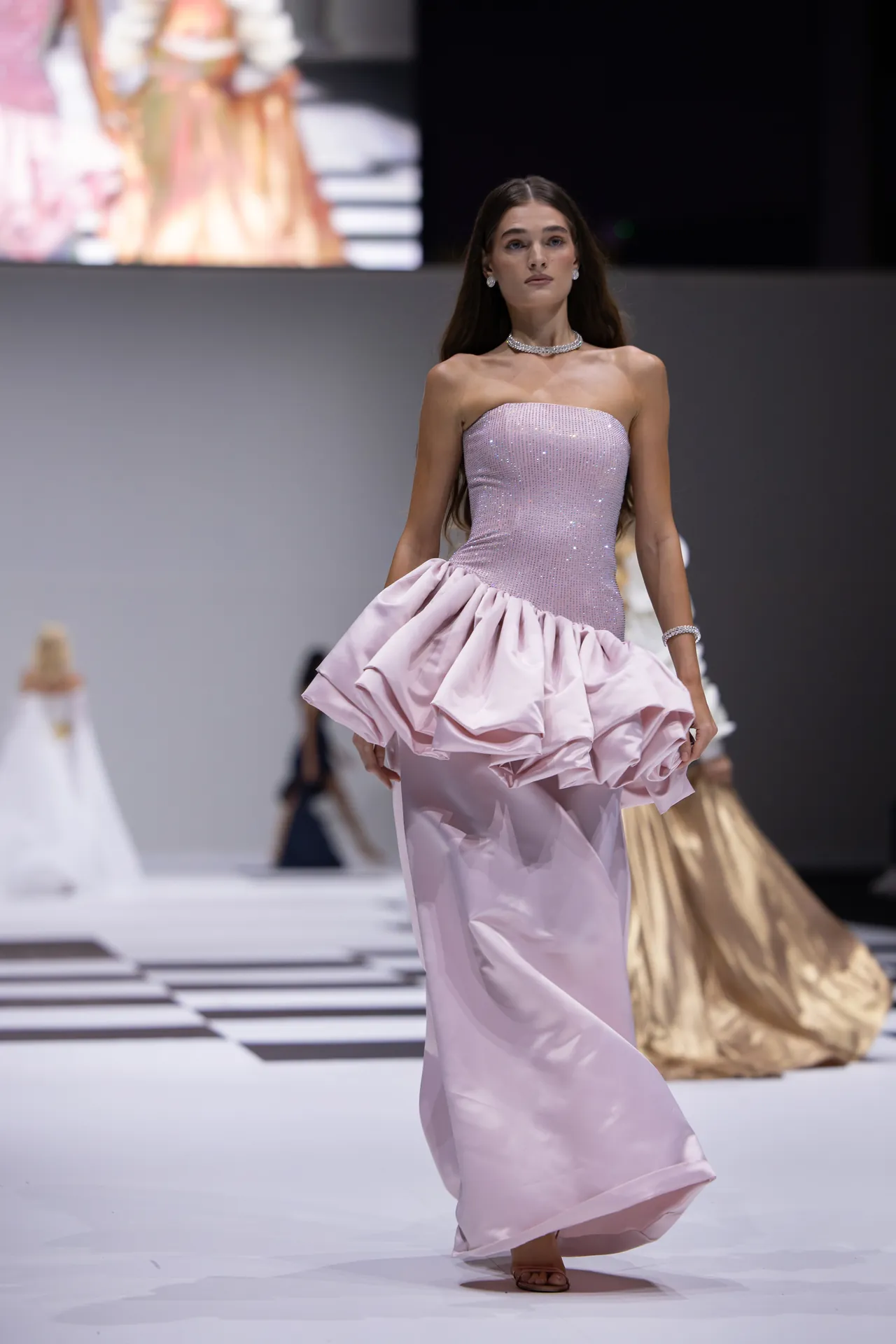Model walking a fashion show in a lavender sequined strapless gown with a large ruffled skirt and jewelry.