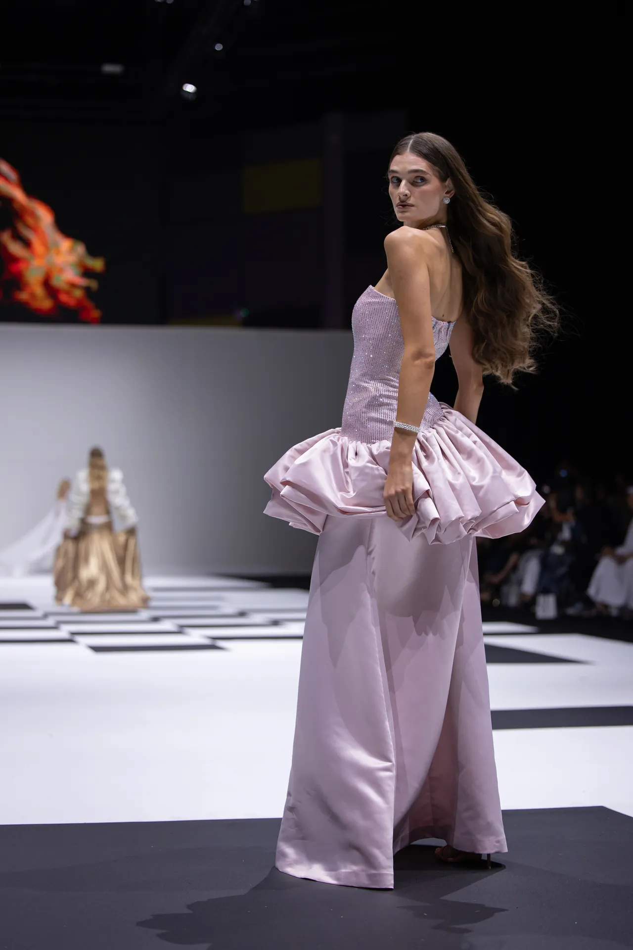 Model walks the runway in a pale pink, strapless satin ballgown with a voluminous, ruffled bustle and sparkling bodice.