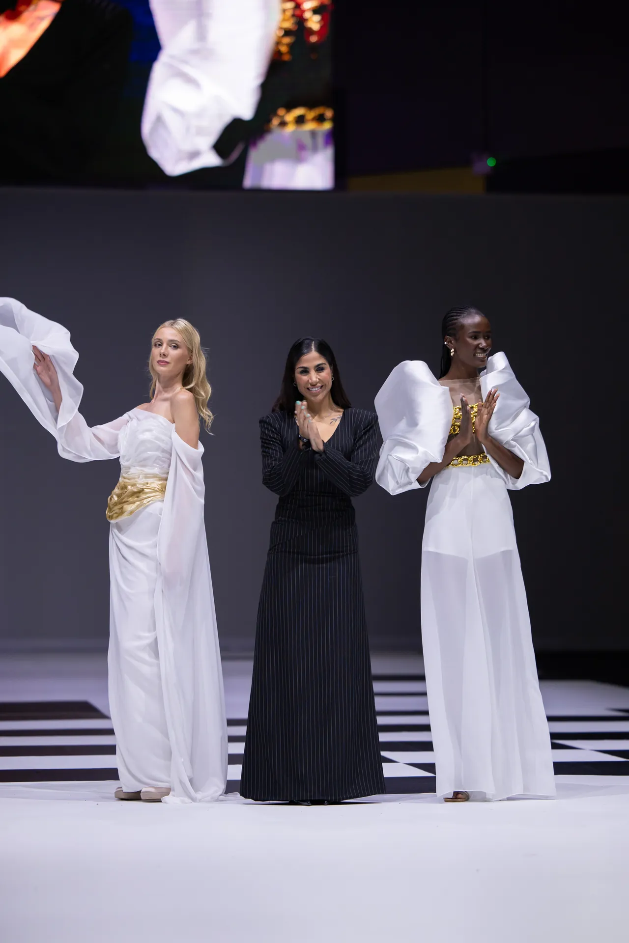 Three women stand on a fashion show runway; two models in white gowns with gold accents flank a woman in a black pinstriped dress, all clapping or bowing on a checkered stage.
