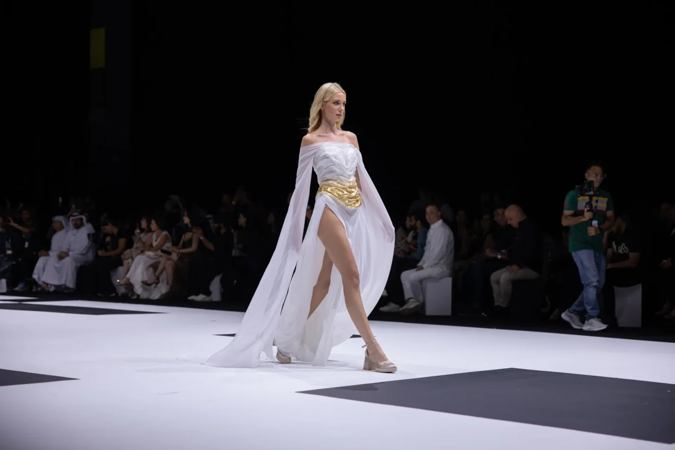 Model walks a white off-shoulder gown with a gold belt and flowing cape on a fashion runway; audience and a cameraman visible in the background.