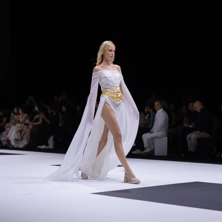 Model walks a white off-shoulder gown with a gold belt and flowing cape on a fashion runway; audience and a cameraman visible in the background.