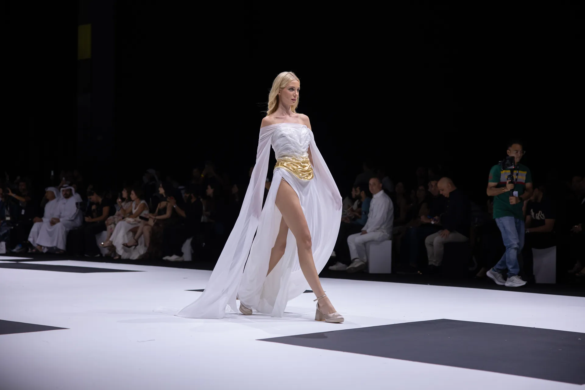 Model walks a white off-shoulder gown with a gold belt and flowing cape on a fashion runway; audience and a cameraman visible in the background.