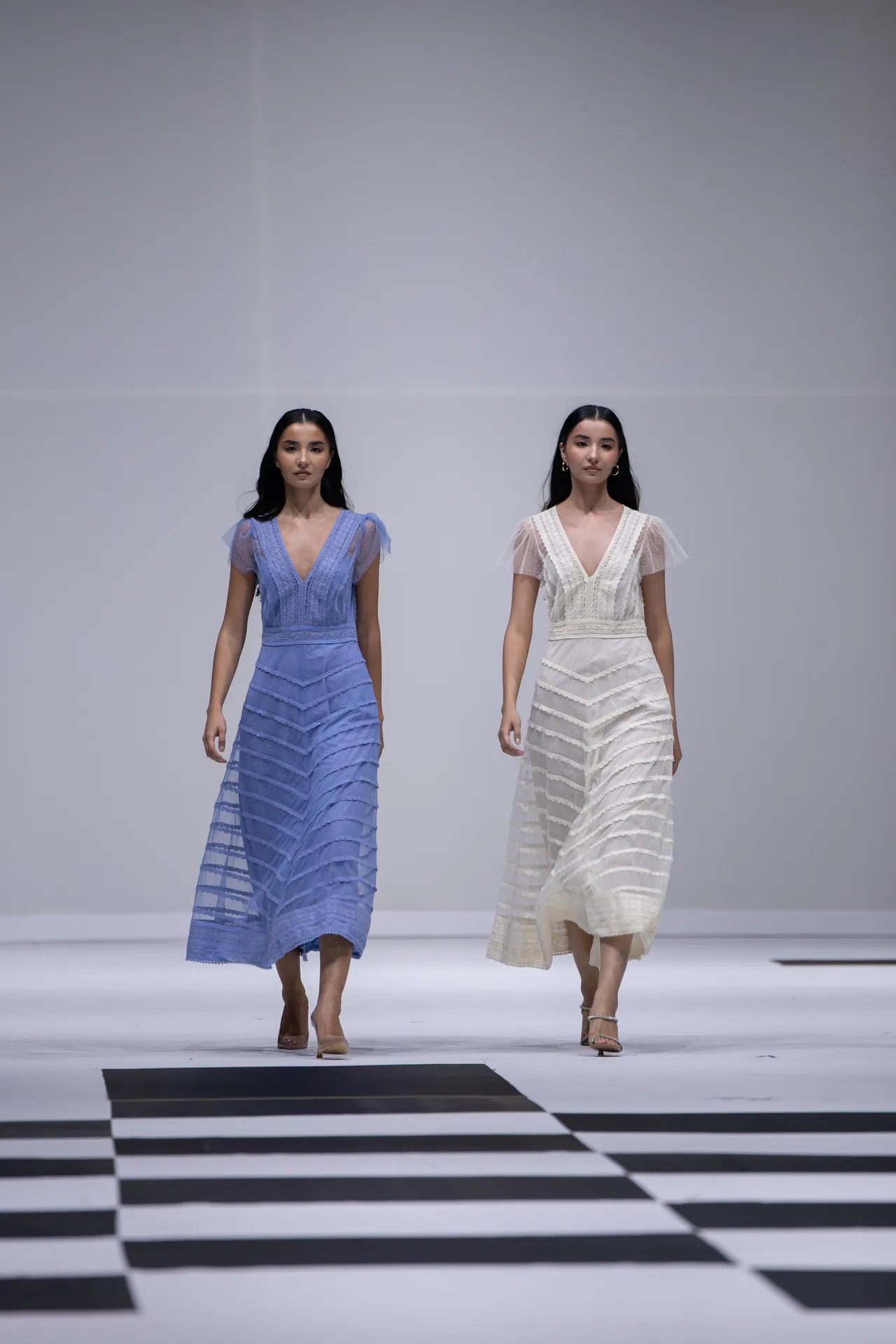 Two models walk a fashion runway in blue and white lace dresses on a black-and-white checkered floor.