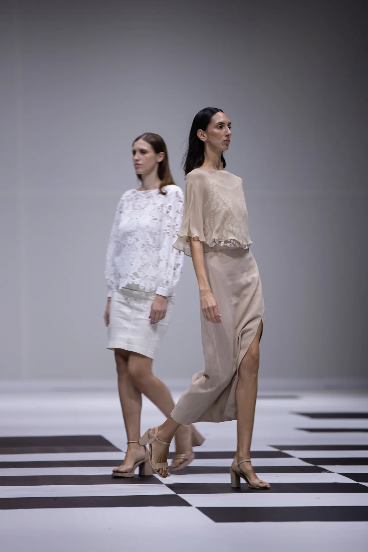 Two female models walk a fashion runway on a black-and-white checkered floor, wearing beige and white lace dresses with heeled sandals.