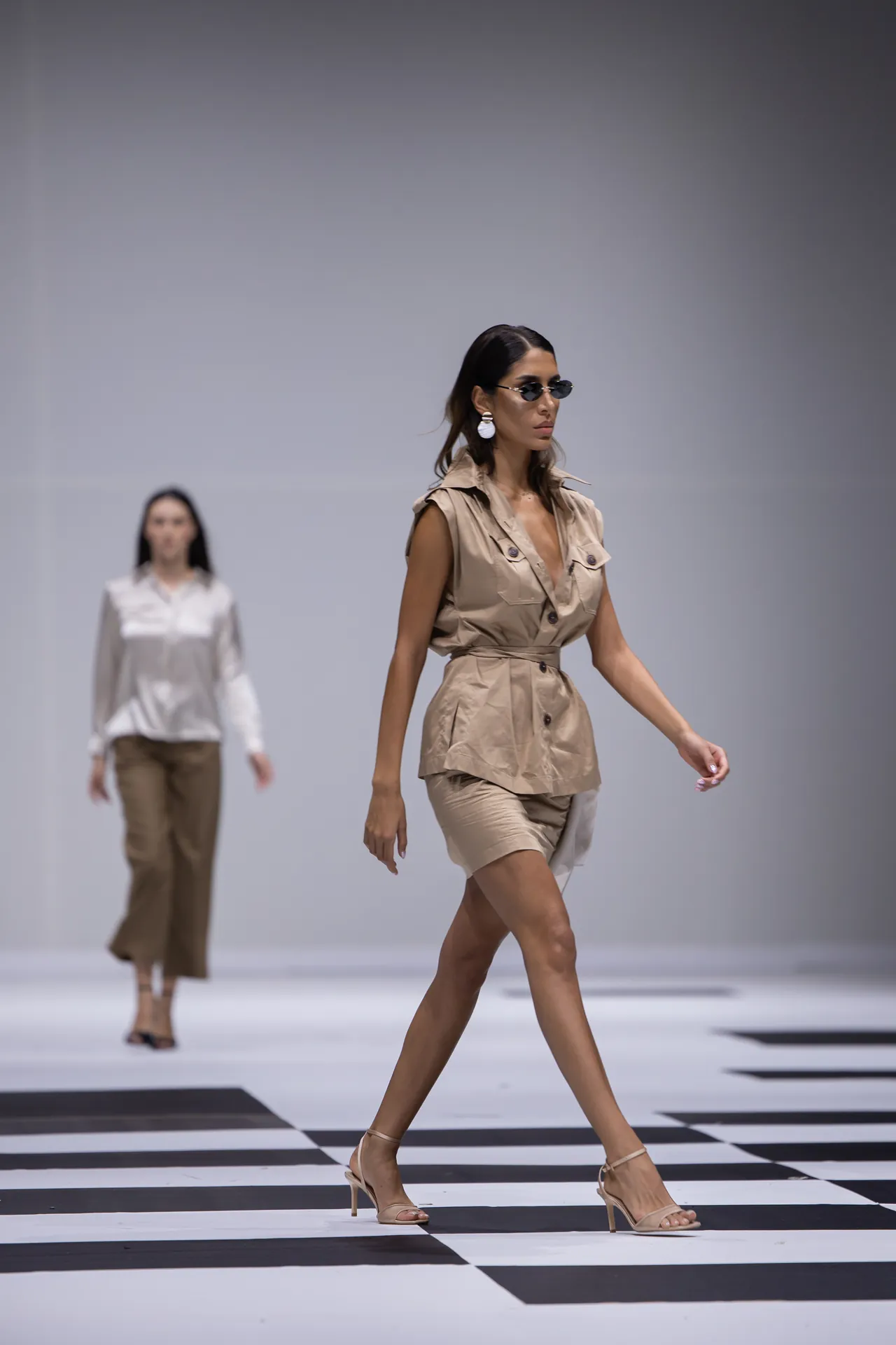 Model walks a fashion runway in a beige sleeveless belted dress, tan heeled sandals, round sunglasses, and white statement earrings on a black-and-white checker floor.