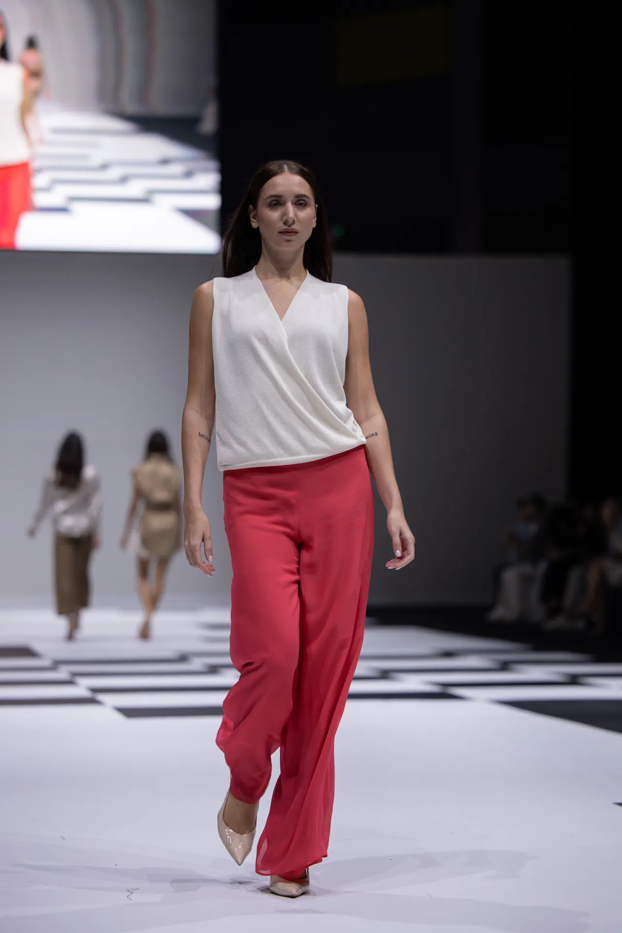 Model walking a fashion runway in a white sleeveless wrap top and red wide-leg pants, beige heels.