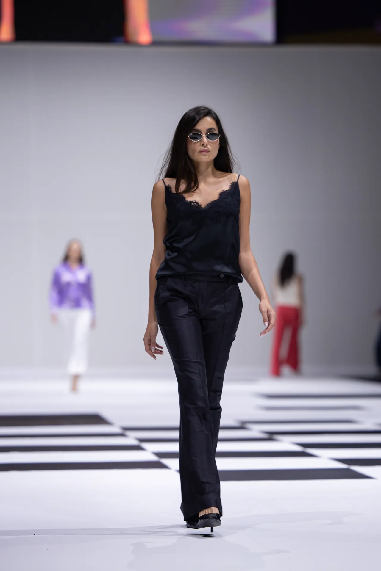 Model walking the runway in a black lace-trim cami and black pants, wearing sunglasses on a checkered catwalk.
